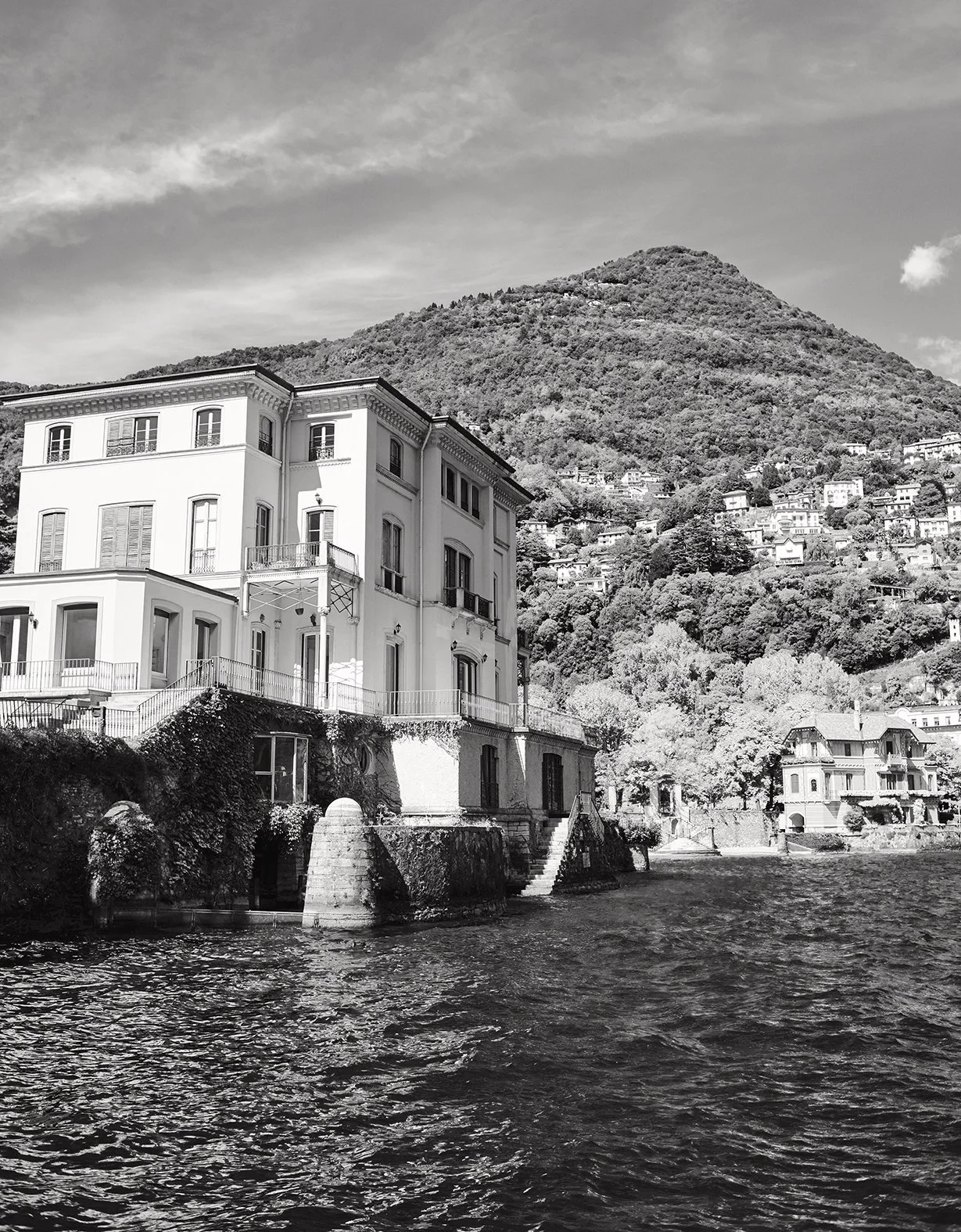 Schwarz-weiß-Foto eines großen Hauses am Wasser mit einer Berglandschaft im Hintergrund.