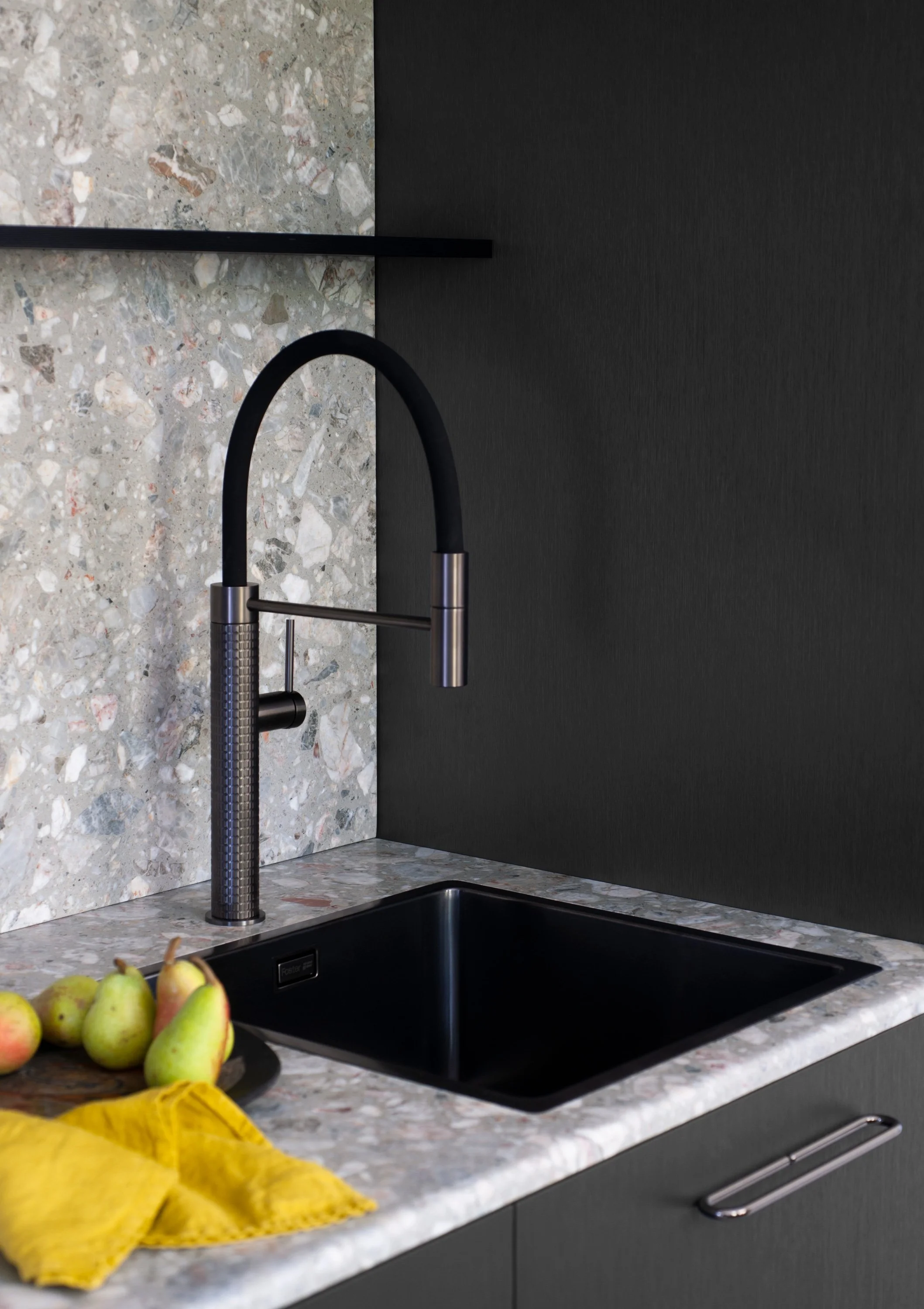 Ein schwarzer Wasserhahn über einer schwarzen Spüle, eingebaut in eine graue Carrara-Marmor-Arbeitsplatte. Im Vordergrund liegt eine gelbe Küchentuch, daneben eine Schüssel mit Birnen auf der Arbeitsfläche. Die Rückwand besteht aus einer schwarzen Wa