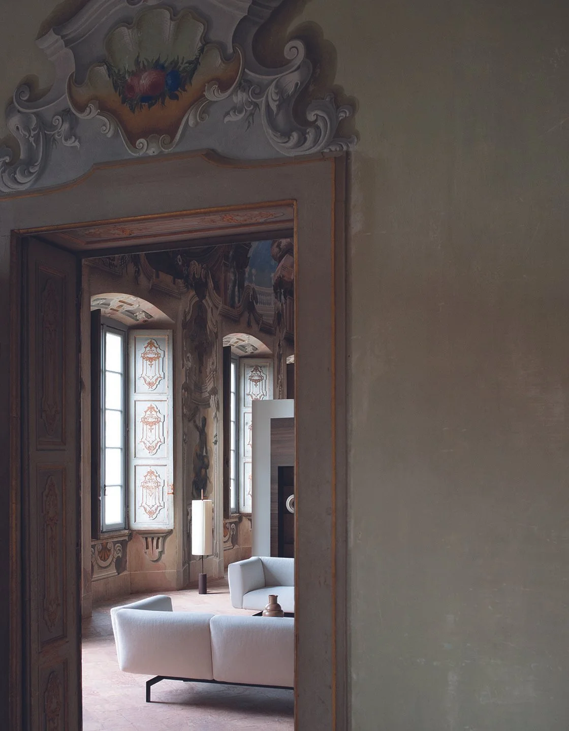 Blick durch eine offene Tür auf einen stilvollen, historischen Raum mit hohen mit barockornamenten verzierten Fenstern und Wandmalereien, moderner weißer Sessel und Lampe.