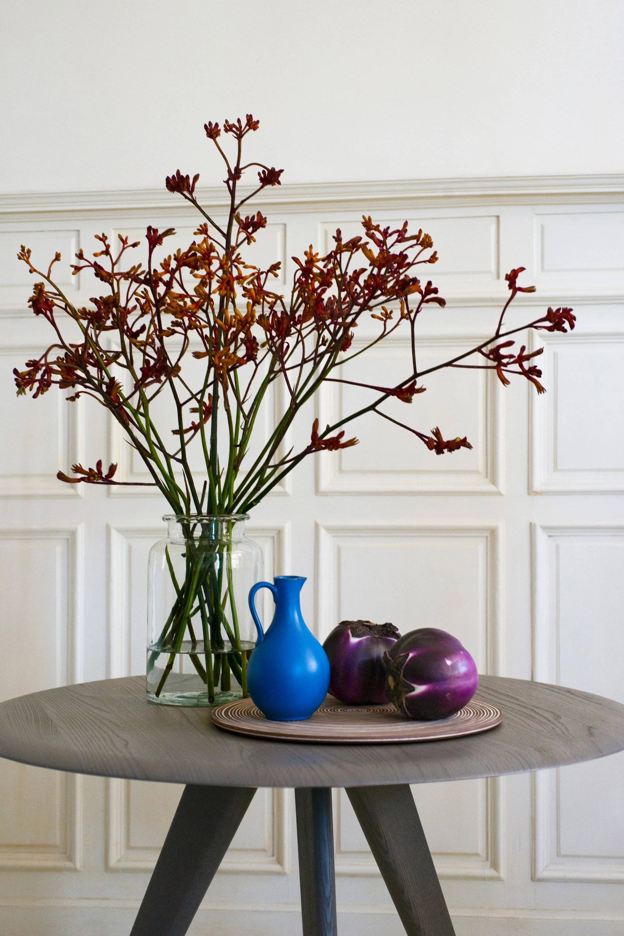 Bunte Blumen in einer Glassvase auf einem Tisch, daneben eine blaue Vase und drei purple Farboteln auf einer runden Holzplatte, im Hintergrund weiße Wand mit Paneelen.