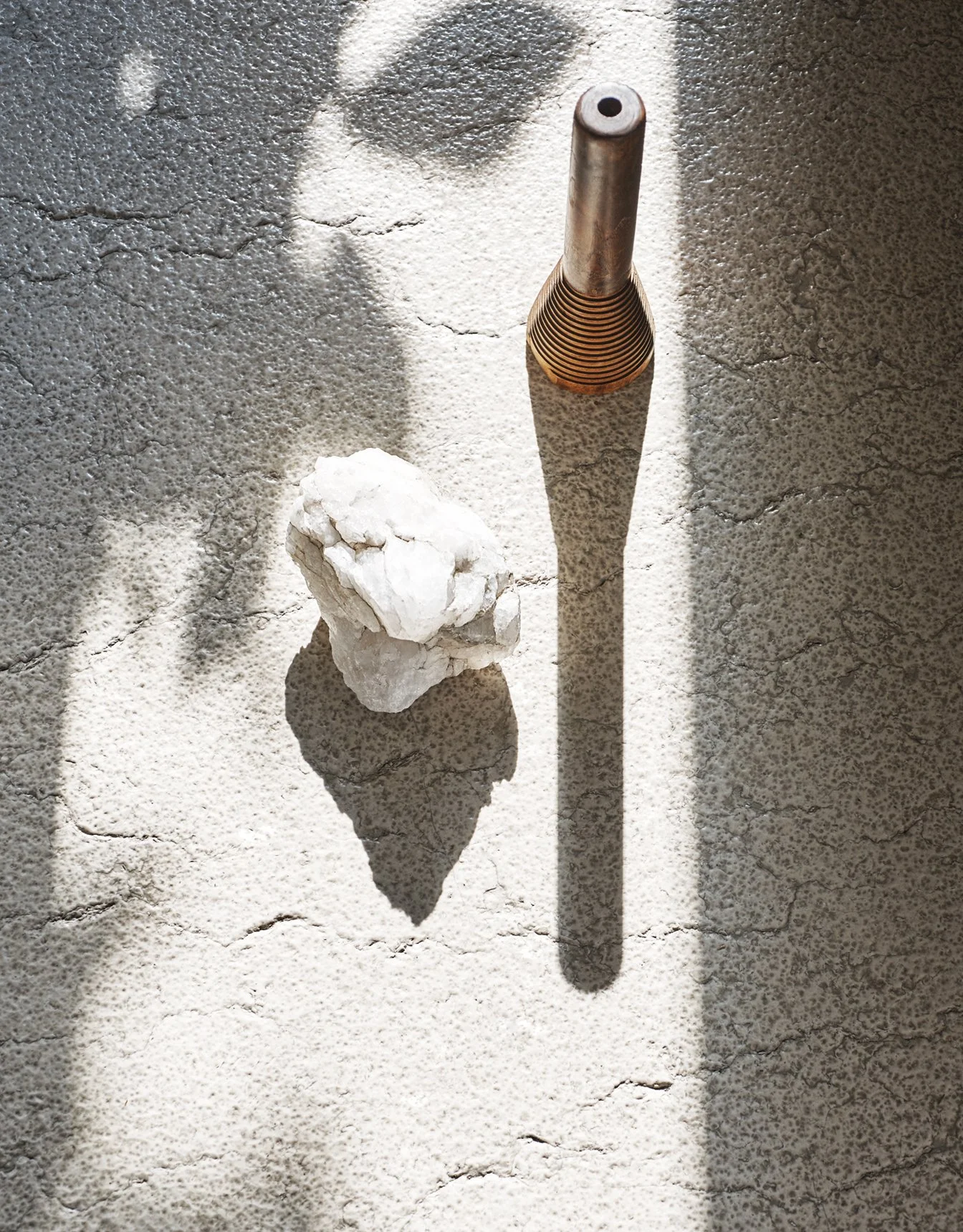 Ein Natursteinboden mit Schattenwurf, auf dem ein kupferfarbener Stab mit einer Spirale und ein weißer Kautzstein liegen.