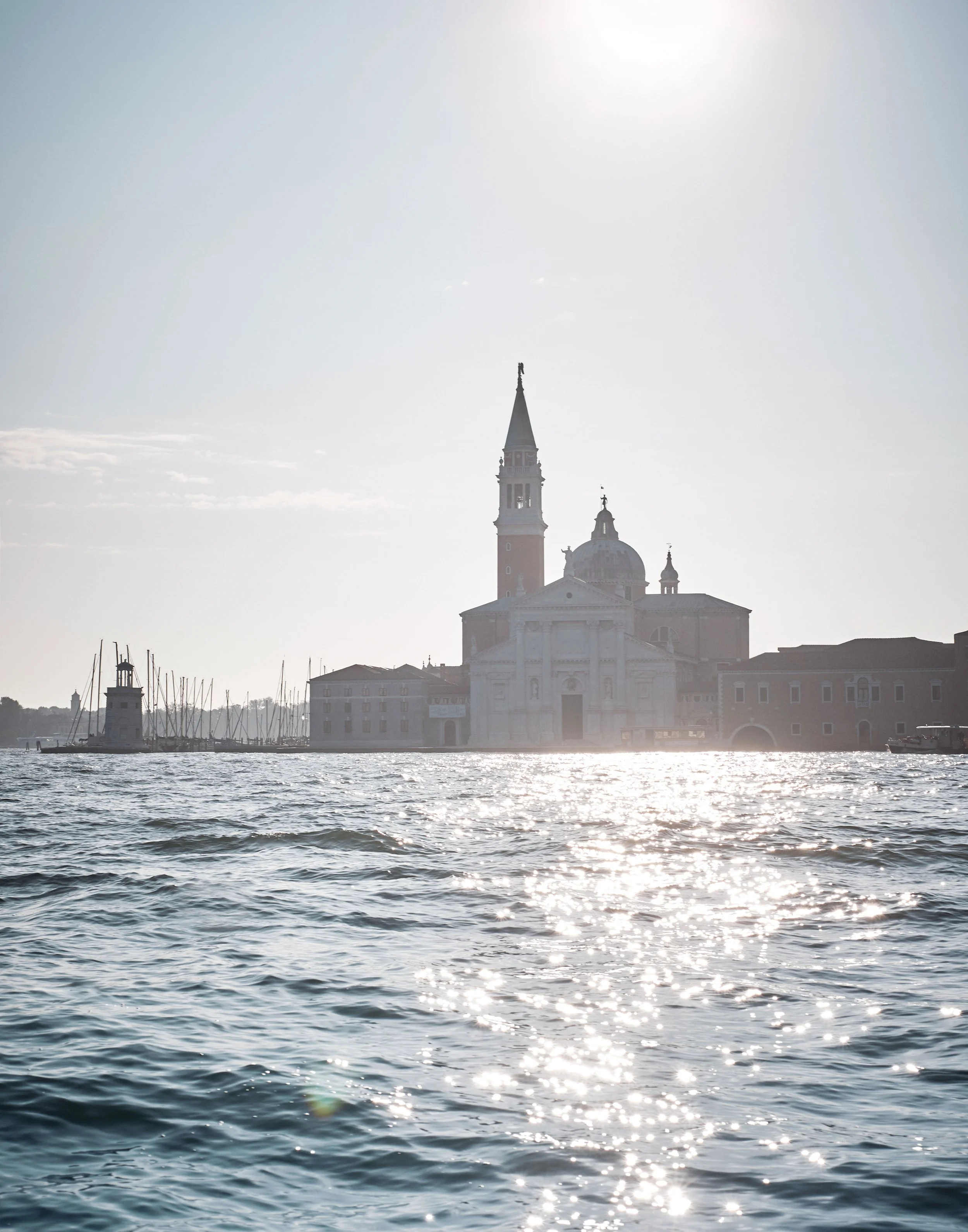 Blick auf das Wasser mit herannahender Sonne und einer Silhouette einer Stadt im Hintergrund, inklusive eines Kirchturms und einiger Gebäude
