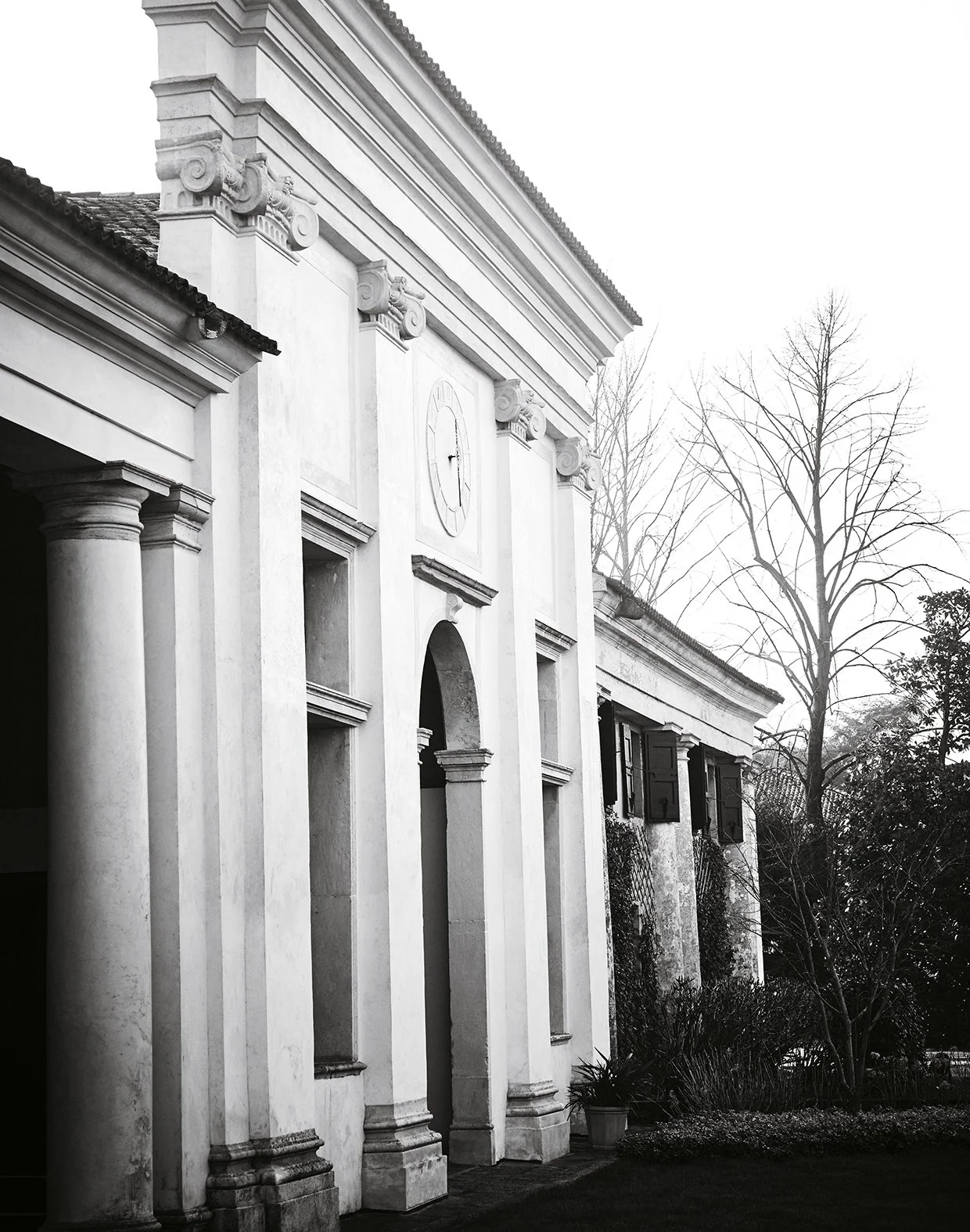 Ein altes, weißes Gebäude mit klassischer Architektur, Fensterläden und einem Uhrenzifferblatt an der Wand. Es gibt Bäume im Hintergrund und einen gepflegten Garten im Vordergrund, alles in Schwarzweiß aufgenommen.