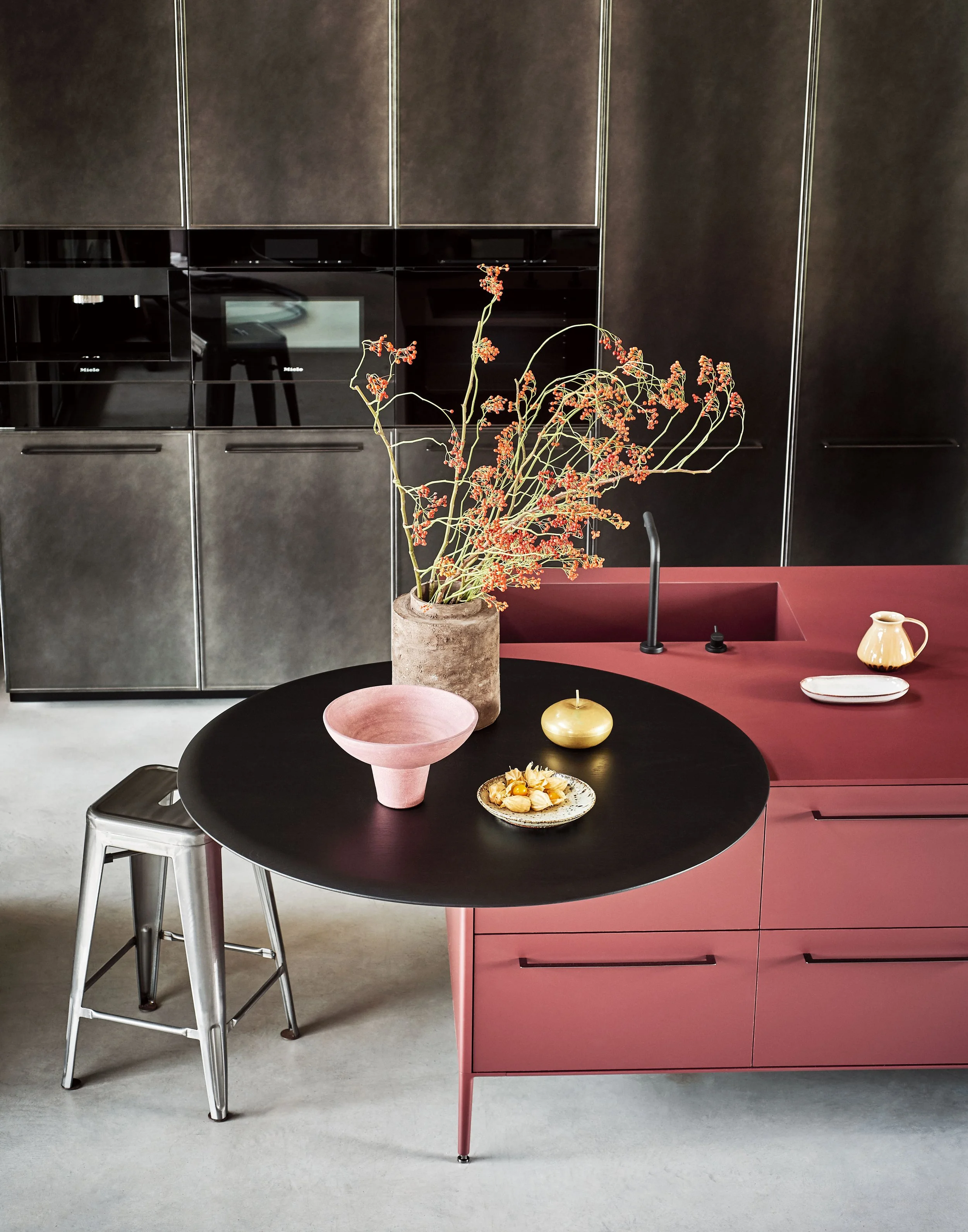 Moderner Küchenraum mit schwarzen und pinkfarbenen Schränken, Vase mit vertrockneten Blumen auf dem Tisch, zwei dekorative Schalen und eine Kanne, schwarze Wasserhähne.