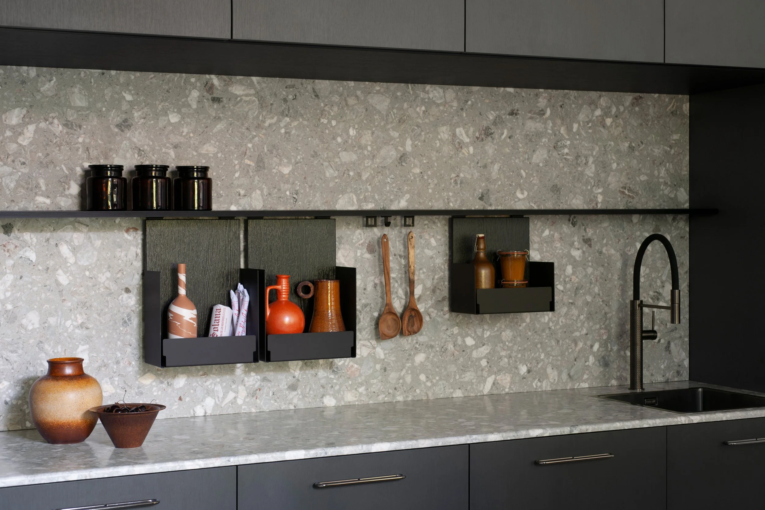 Moderne Küchenarbeitsplatte aus grauem Stein mit schwarzen Wandregalen, Keramikvasen in Erdtönen, Holzlöffeln, Küchenutensilien und dunklen Schränken.