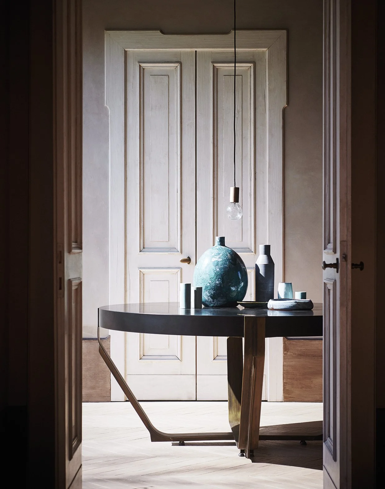 Ein Raum mit doppelten Holztüren im Hintergrund, auf einem runden Tisch stehen mehrere Vasen und Keramikobjekte, darunter eine große blaue Vase, eine schwarze und mehrere kleine weiße Vasen, Licht fällt durch eine unsichtbare Lichtquelle, schafft ein