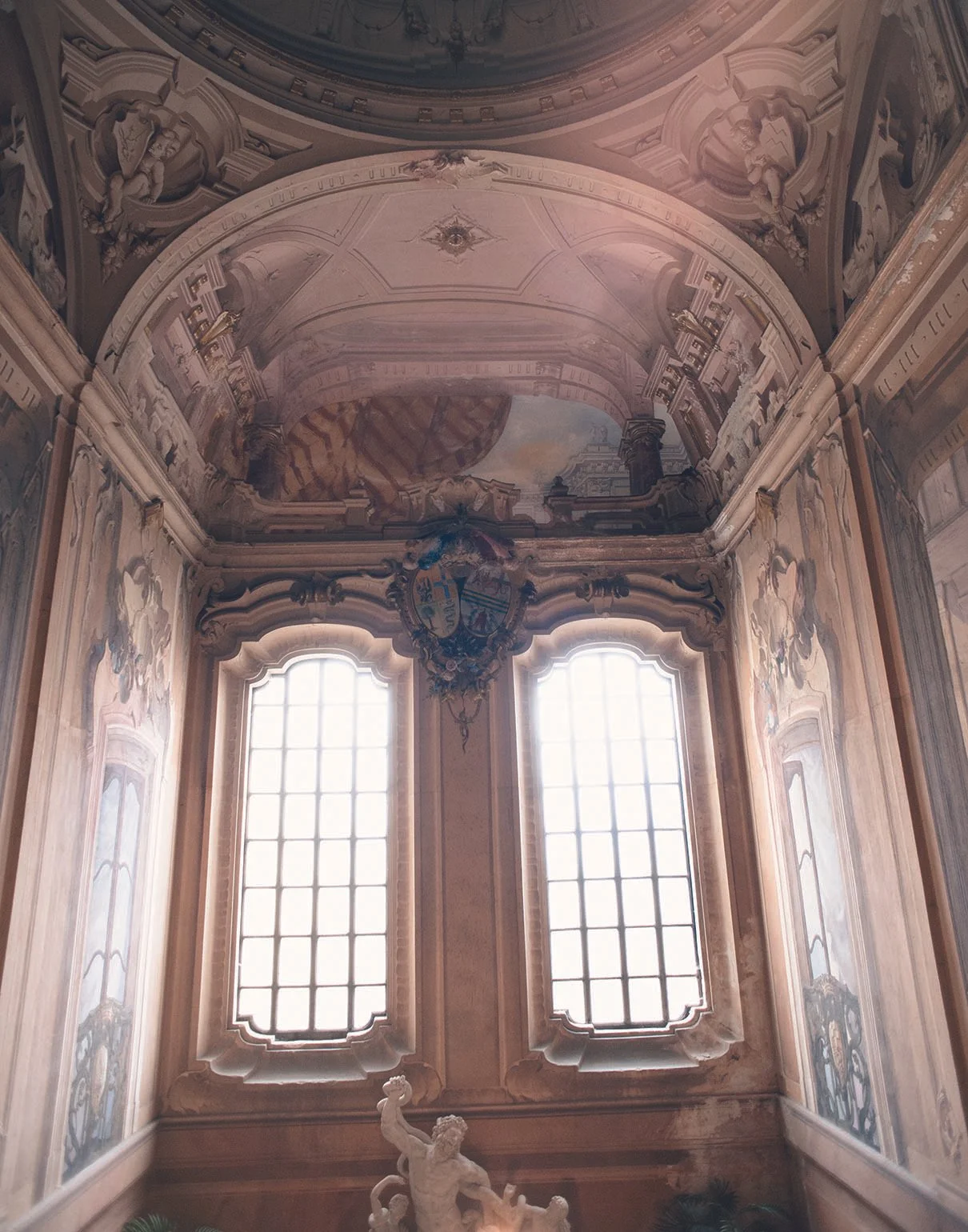 Ein Barocksaal mit hohen Fenstern, kunstvollen Deckenmalereien und einer Skulptur im Vordergrund.