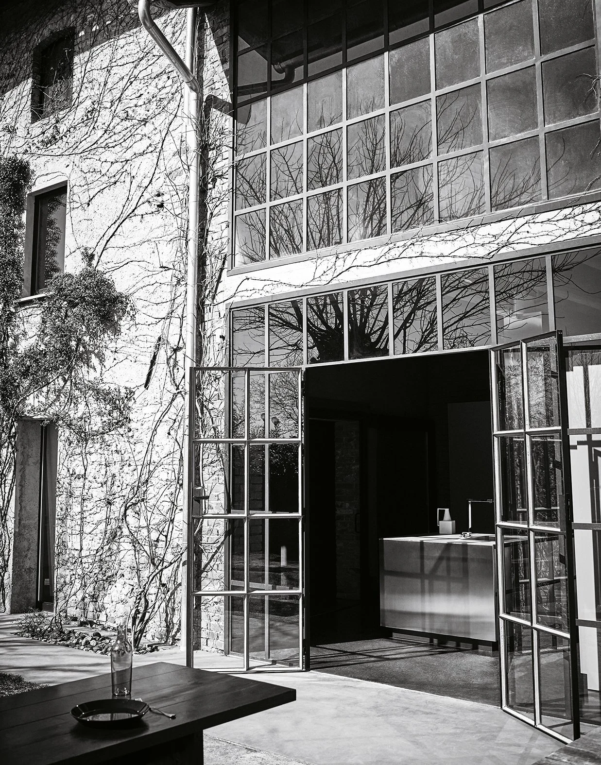 Ein moderner Garten mit großen Fensterflächen und einem offenen Eingang, im Hintergrund sind kahle Bäume sichtbar, die auf die Jahreszeit Frühling oder Winter hinweisen.