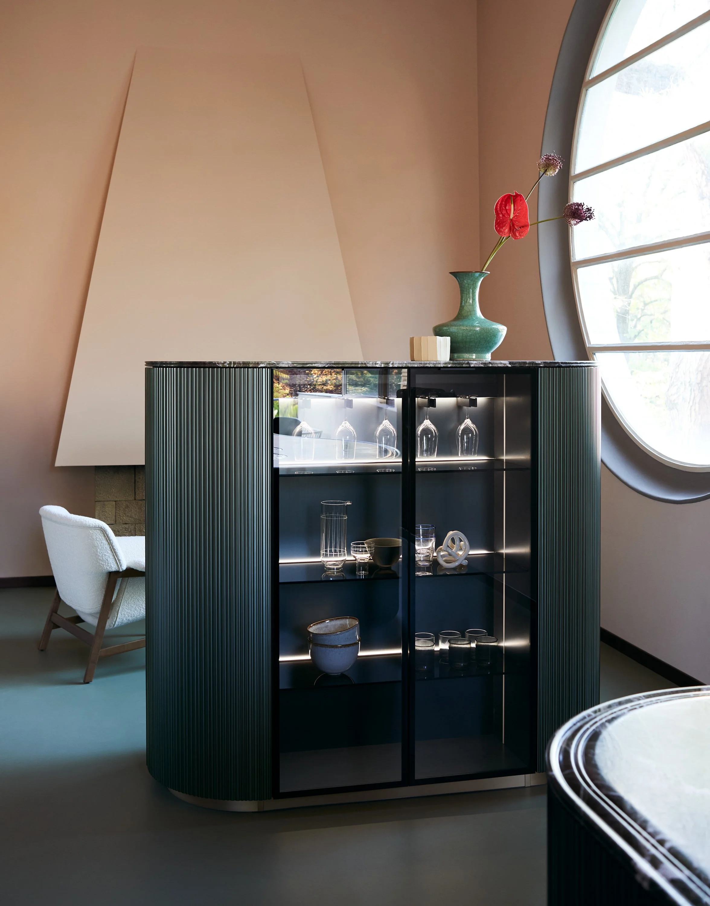 Ein stilvoller, moderner Raum mit einer schwarzen Vitrine, einem grünen Keramikvase mit roten und lila Blumen auf der Topfplatte, ein weißes Sitzmöbel auf der linken Seite und einer großen Fenster mit elliptischer Form auf der rechten Seite.