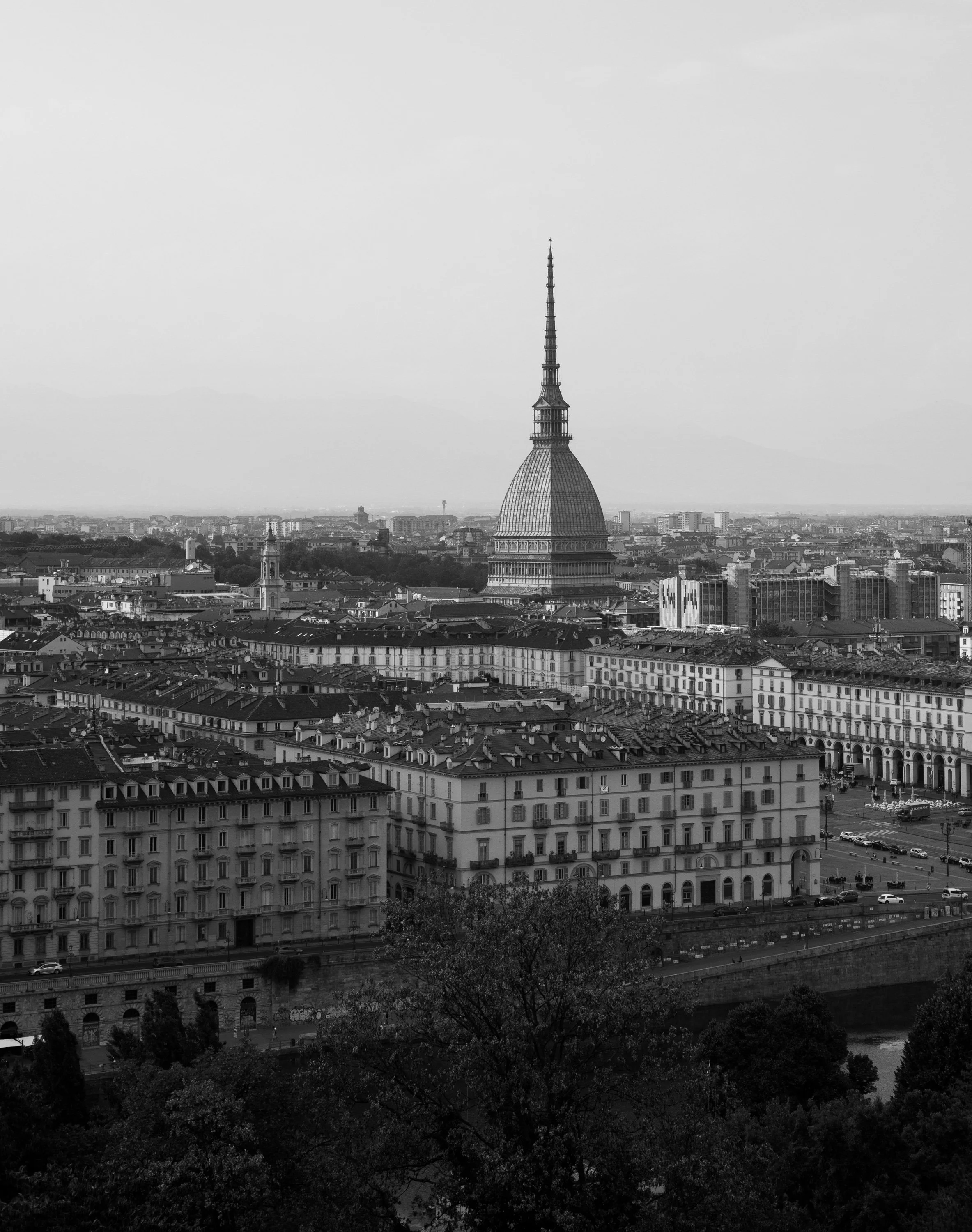 Schwarz-weiß-Fotografie einer Stadt mit einer markanten Turmspitze, vermutlich die Mole Antonelliana in Turin, im Hintergrund Berge und ein Wolkenhimmel.