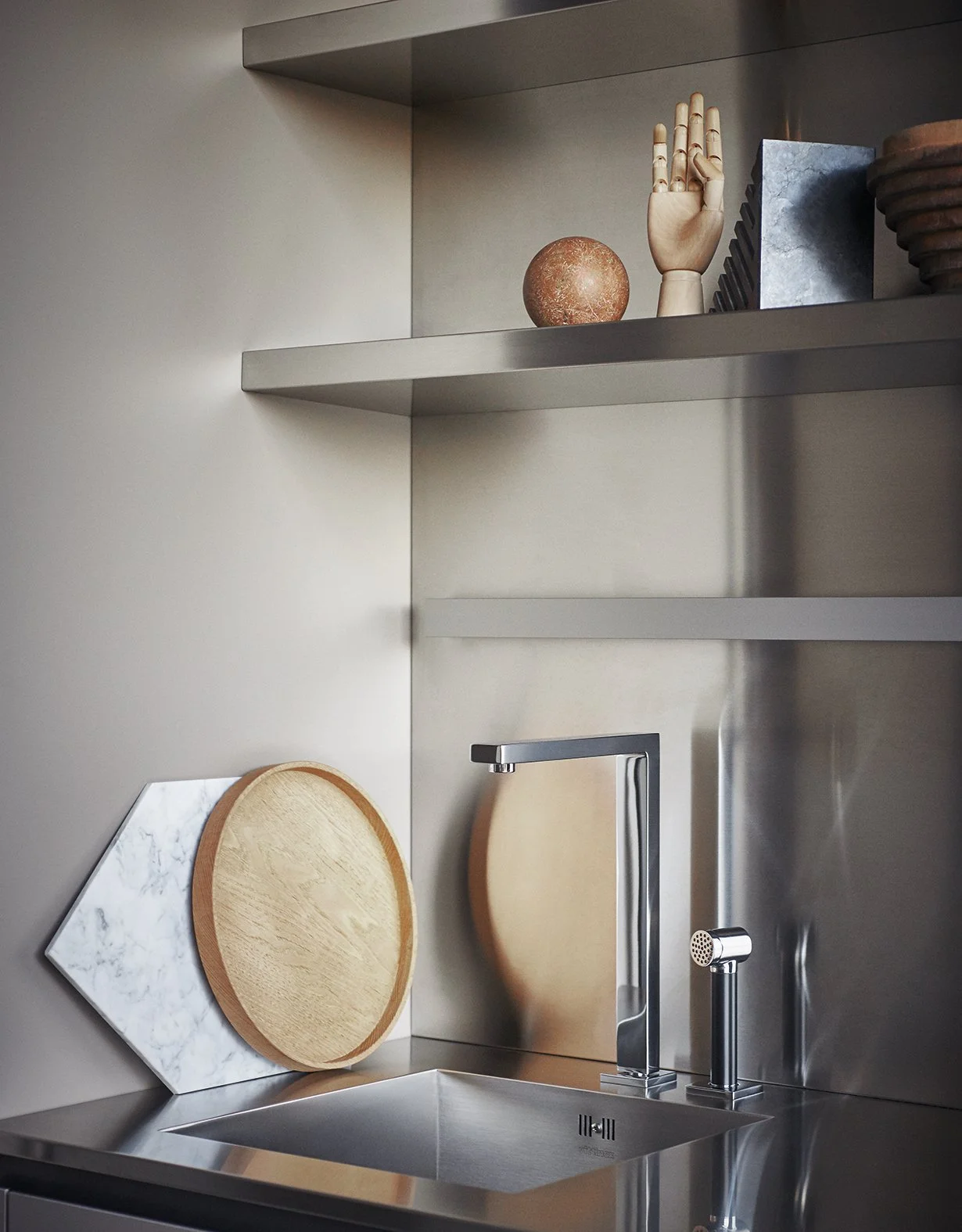 Moderne Küchenspüle mit Edelstahl-Armatur, zwei runde Schneidebretter und dekorative Gegenstände auf den Regalen, darunter eine Holzhand, eine kleine Kugel, Steine und Keramikplatten.