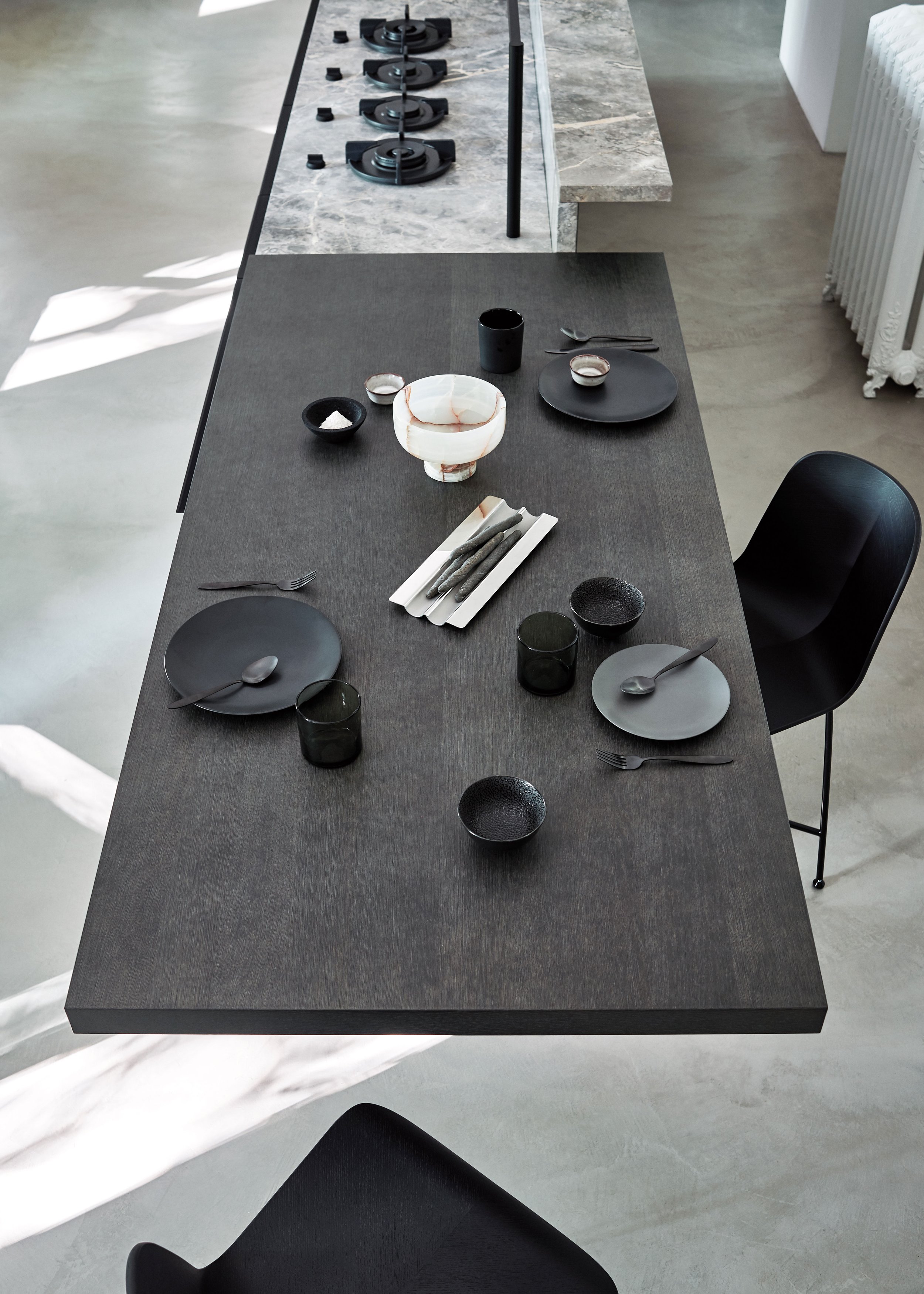 Ein moderner Esstisch mit schwarzen und weißen Geschirr, Besteck, dunklen Trinkgläsern und einer schwarzen Tasse, in einer hell erleuchteten, minimalistischen Küche mit glattem Boden und einem Herd im Hintergrund.