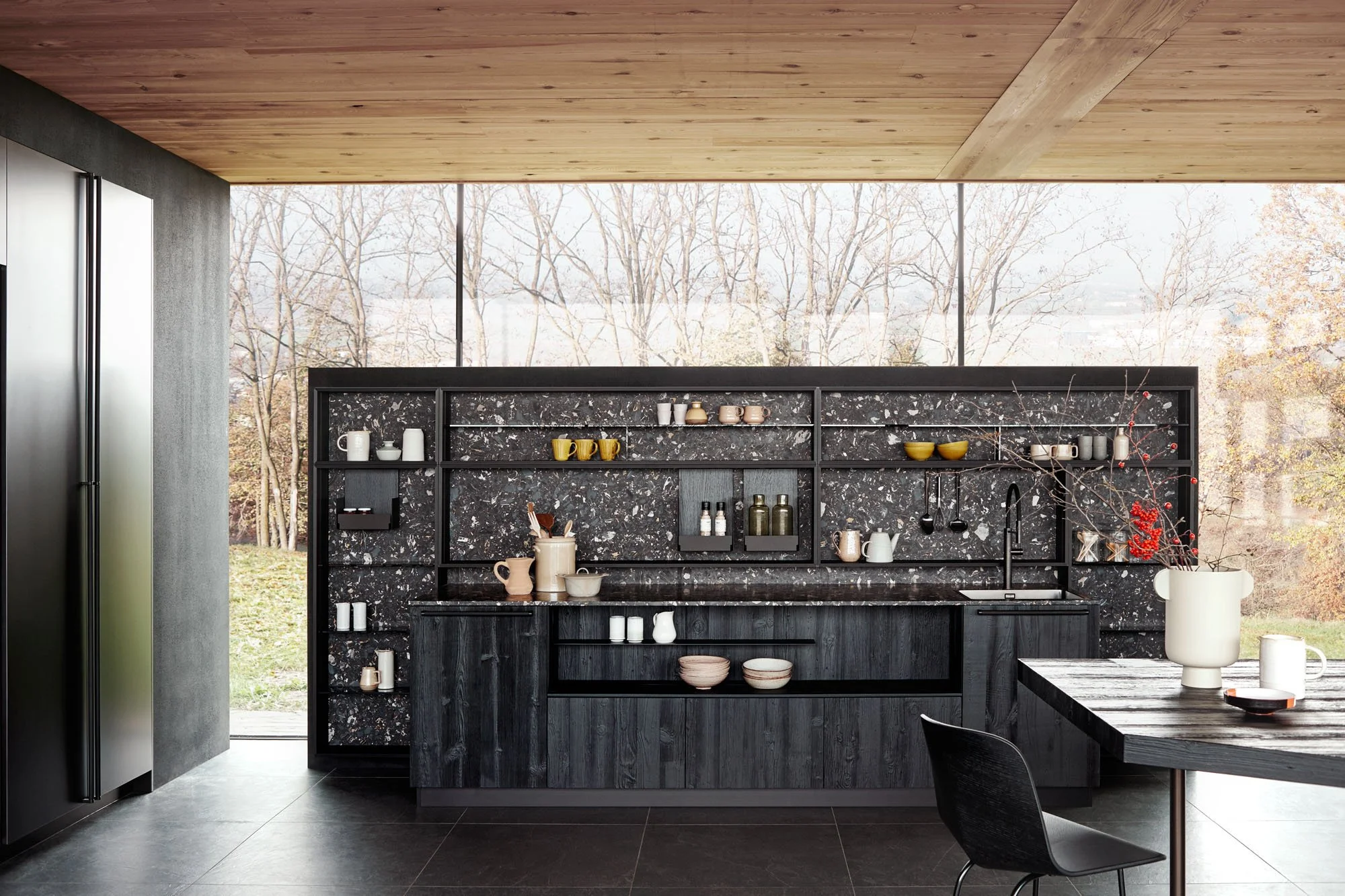 Moderne schwarze Küchenzeile mit grauem und schwarzem Marmor, offene Regale, diverse Tassen, Schalen und Dekorationen, große Fensterscheibe mit Blick auf Bäume im Winter, Holzdecke, grauer Boden, schwarzer Esstisch mit Stuhl im Vordergrund.