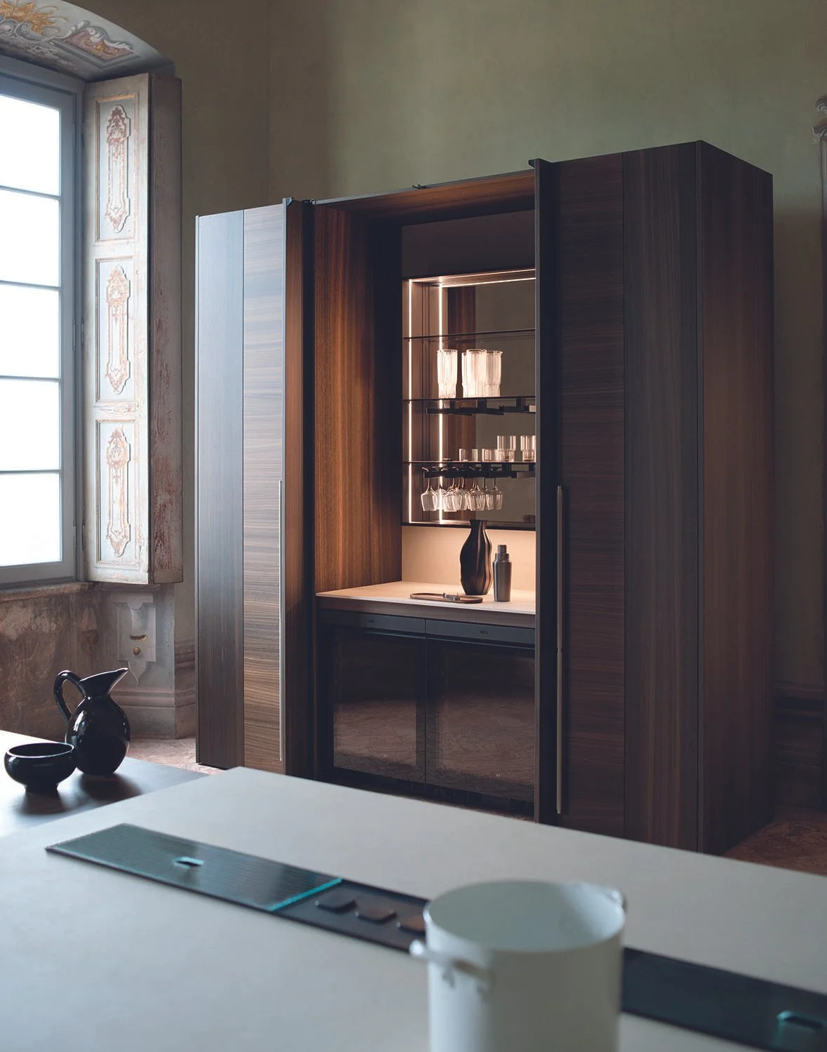 Hinteres Schlafzimmer mit einem modernen, dunklen Holzschrank und Glasregalen, die mit Gläsern und Vasen dekoriert sind, neben einem großen Fenster mit aufwändiger Rahmenverzierung.