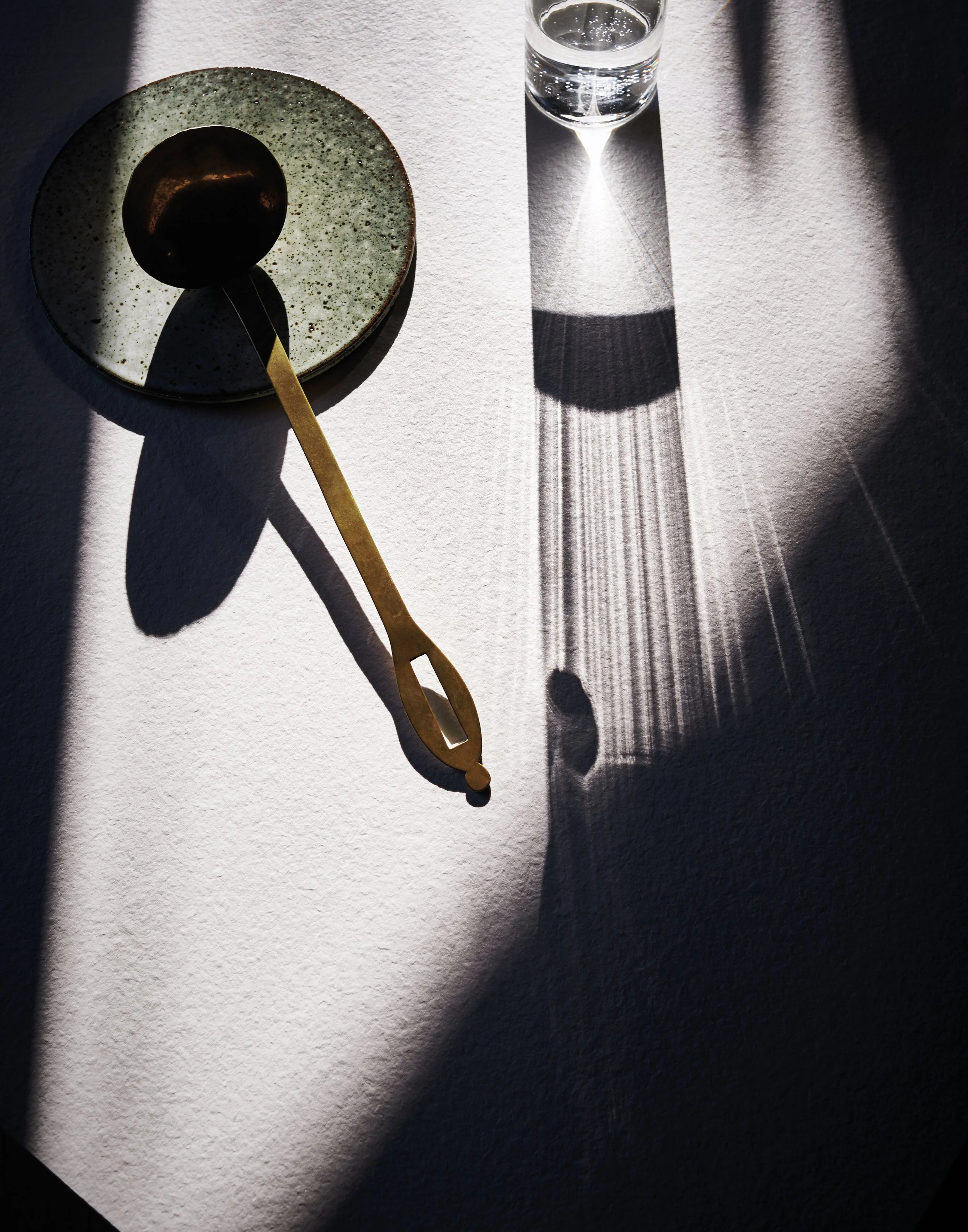 Eine kleine grüne Schale mit dunklem Gegenstand darin, ein goldener Löffel, eine aufgekippte Wasserflasche, alles auf einem hellen Tisch. Sonnenlicht wirft Schatten, die bestimmte Gegenstände auf der Oberfläche betonen.