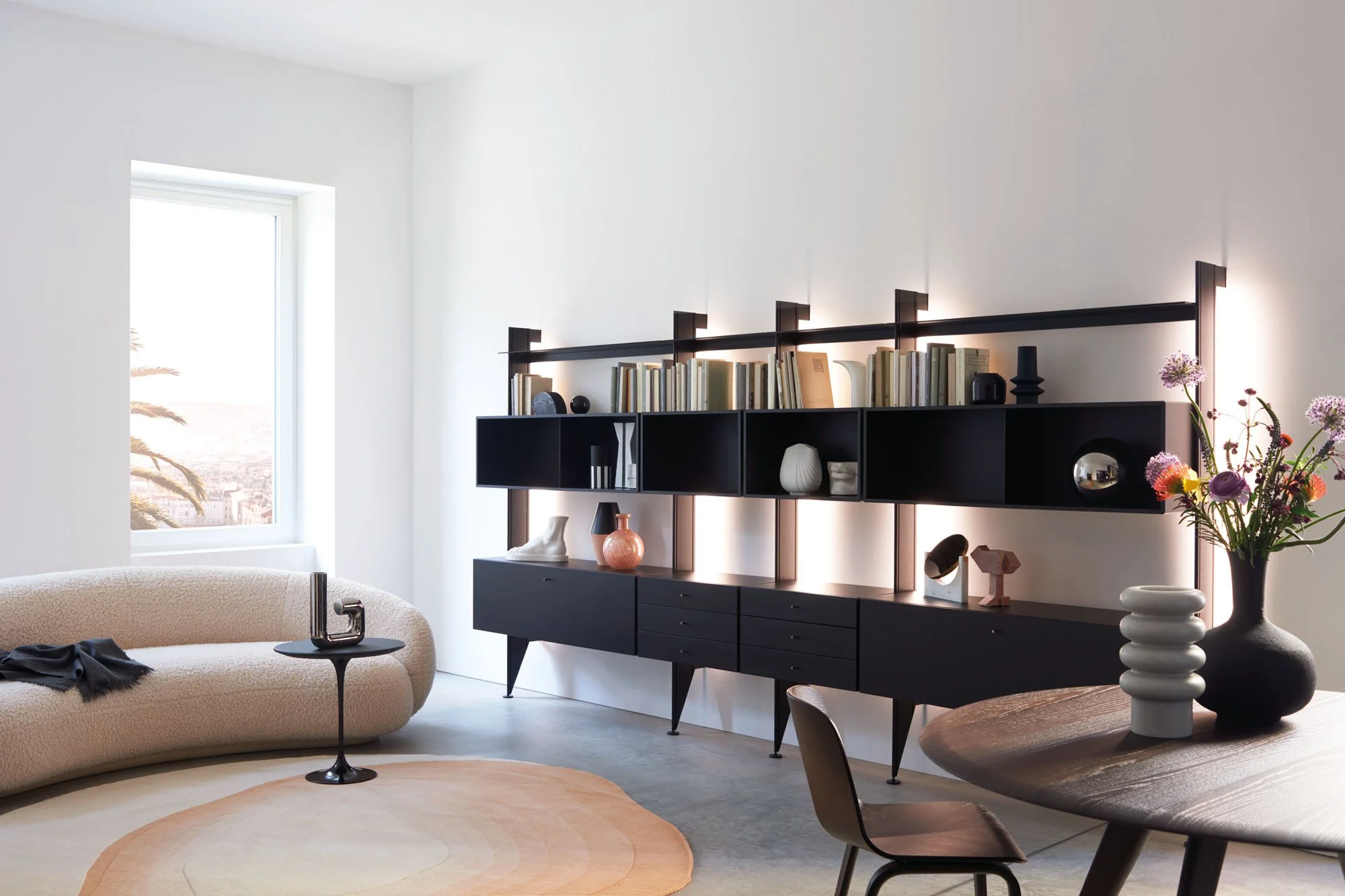 Modernes Wohnzimmer mit schwarzen Regalen, weißen Wänden, Pflanzen und Dekorationsobjekten, große Fenster mit Stadtblick.