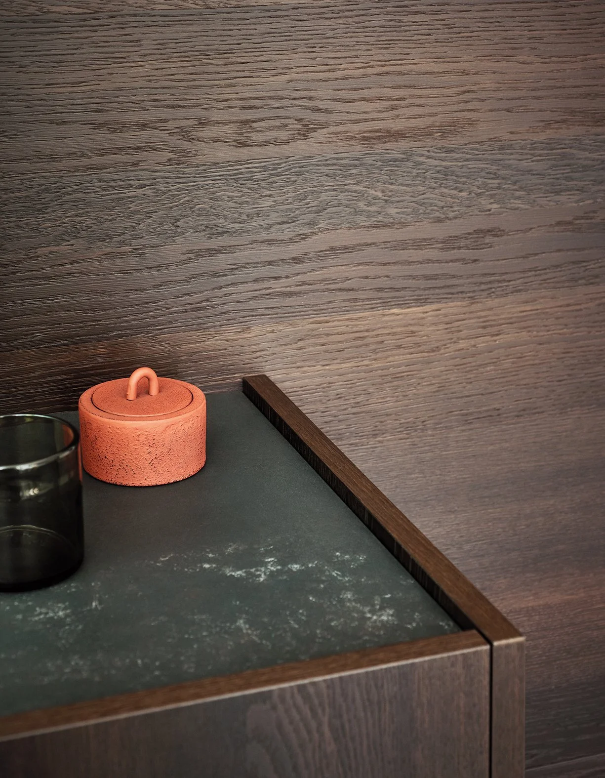 Ein schwarzer Tisch mit Staub und einer kleinen Tasche mit Griffen, daneben ein dunkles Glas und eine Wand mit Holzoptik im Hintergrund.