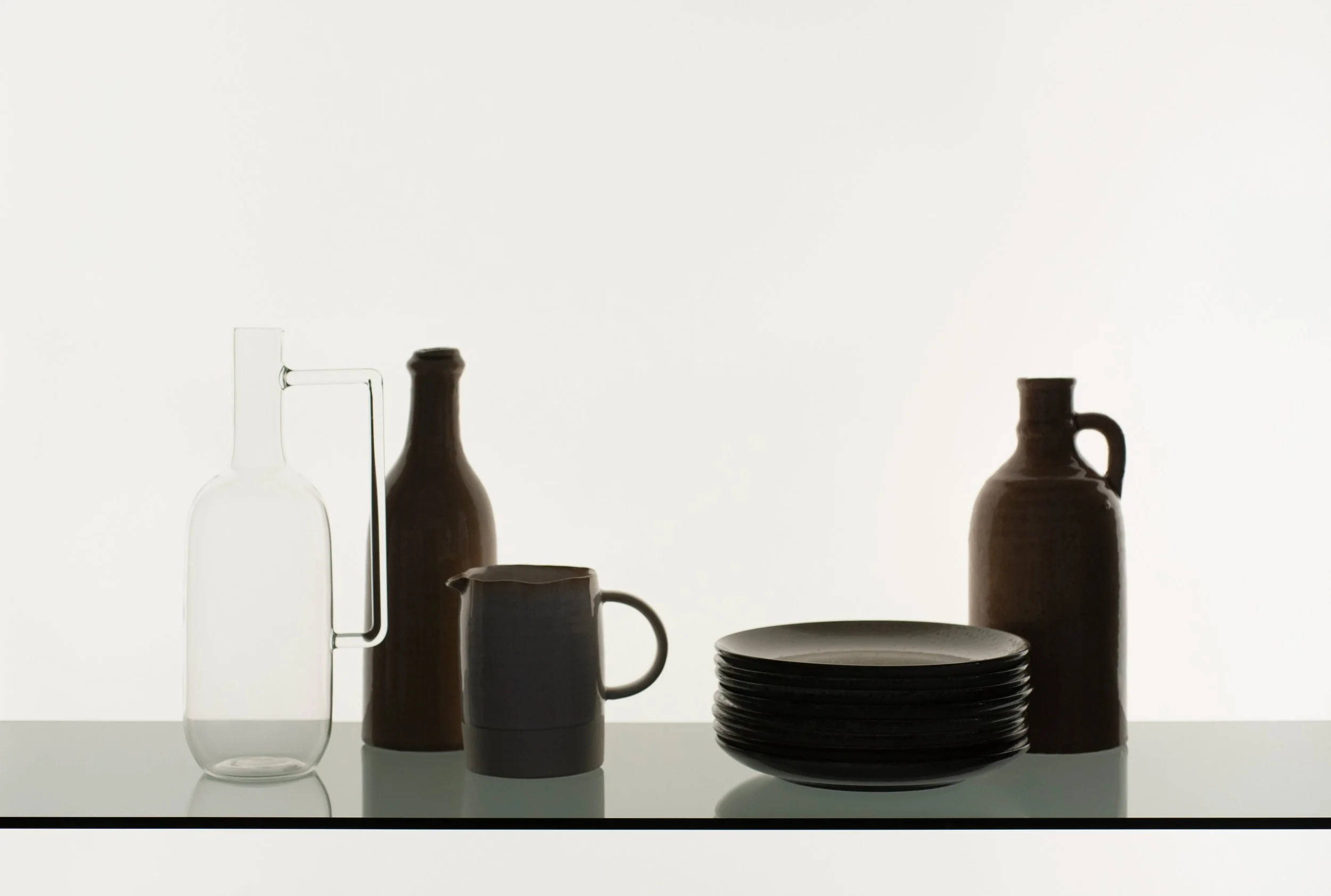 Transparente Glasflasche, braune Flasche, braune Kaffeekanne, graue Tasse, stapel schwarze Teller, braune kleine Flasche mit Griff, alle Gegenstände auf einer weißen Oberfläche vor hellem Hintergrund.