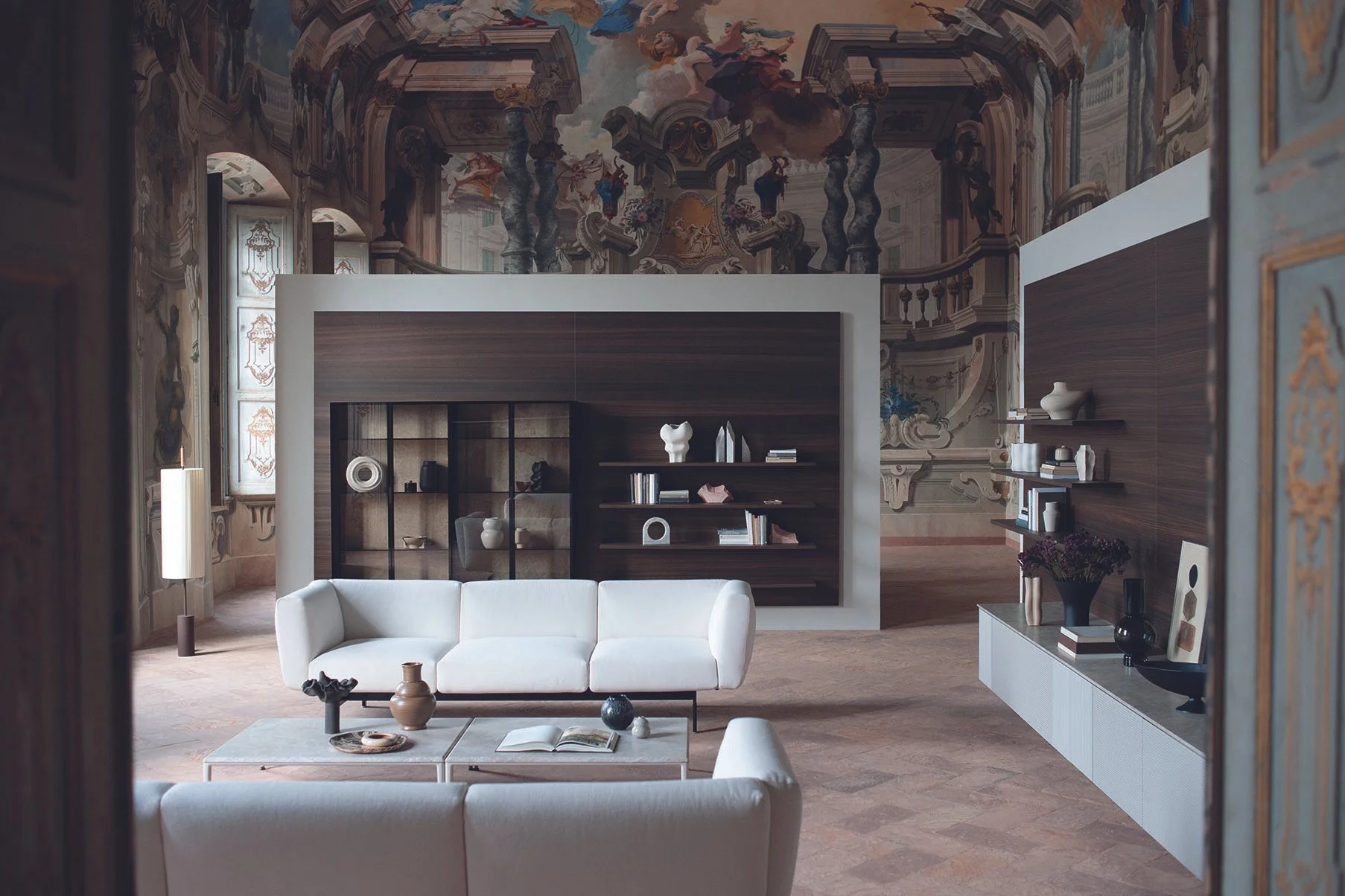 Innenansicht eines eleganten Wohnzimmers mit Barockdekor an der Decke, moderner Möbeln, weißen Sofas, dunklen Holzregalen und Dekorationsartikeln wie Vasen, Büchern und Skulpturen.