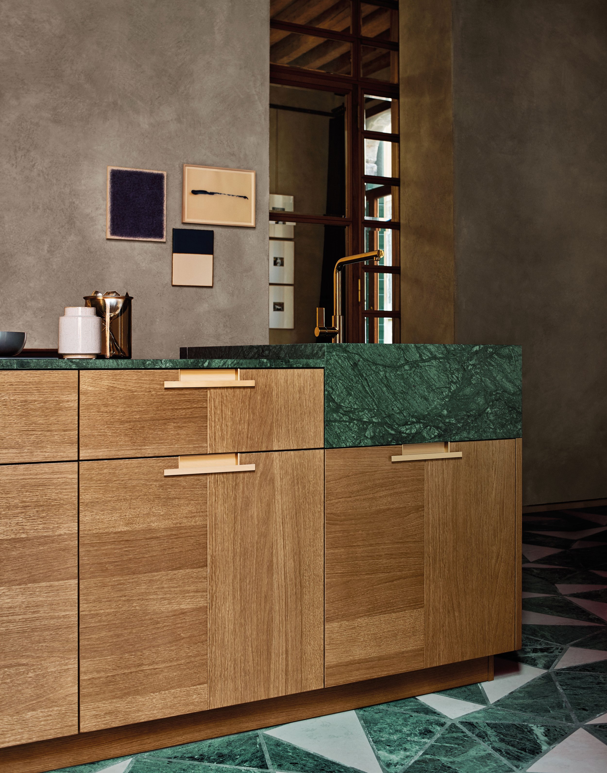 Ein Küchenblock mit Holzschränken, einer grünen Marmor-Arbeitsplatte und einem schwarzen Wasserhahn. Im Hintergrund sind Wandbilder und eine Wand mit Fenstern zu sehen.
