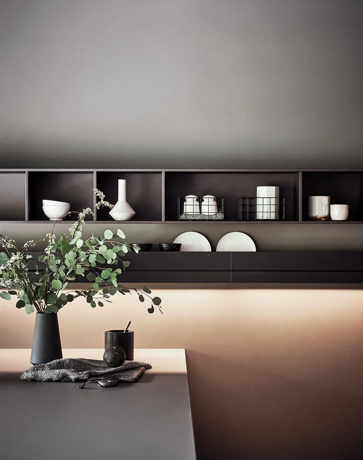Hintergrundwandschrank mit weißen Keramikvasen, kleinen Schalen und dekorativen Fässern, und im Vordergrund ein Tisch mit einer Vase mit Grünpflanzen, einer schwarzen Kerze, einem dunklen Stein, einem Messer, einem Stoff und einem Becher.