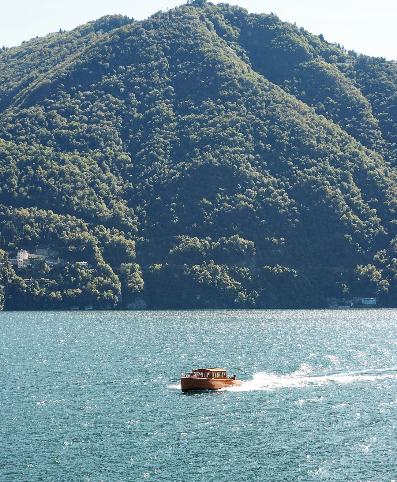 Ein Holzboot fährt auf einem See mit grünen, bewaldeten Bergen im Hintergrund.