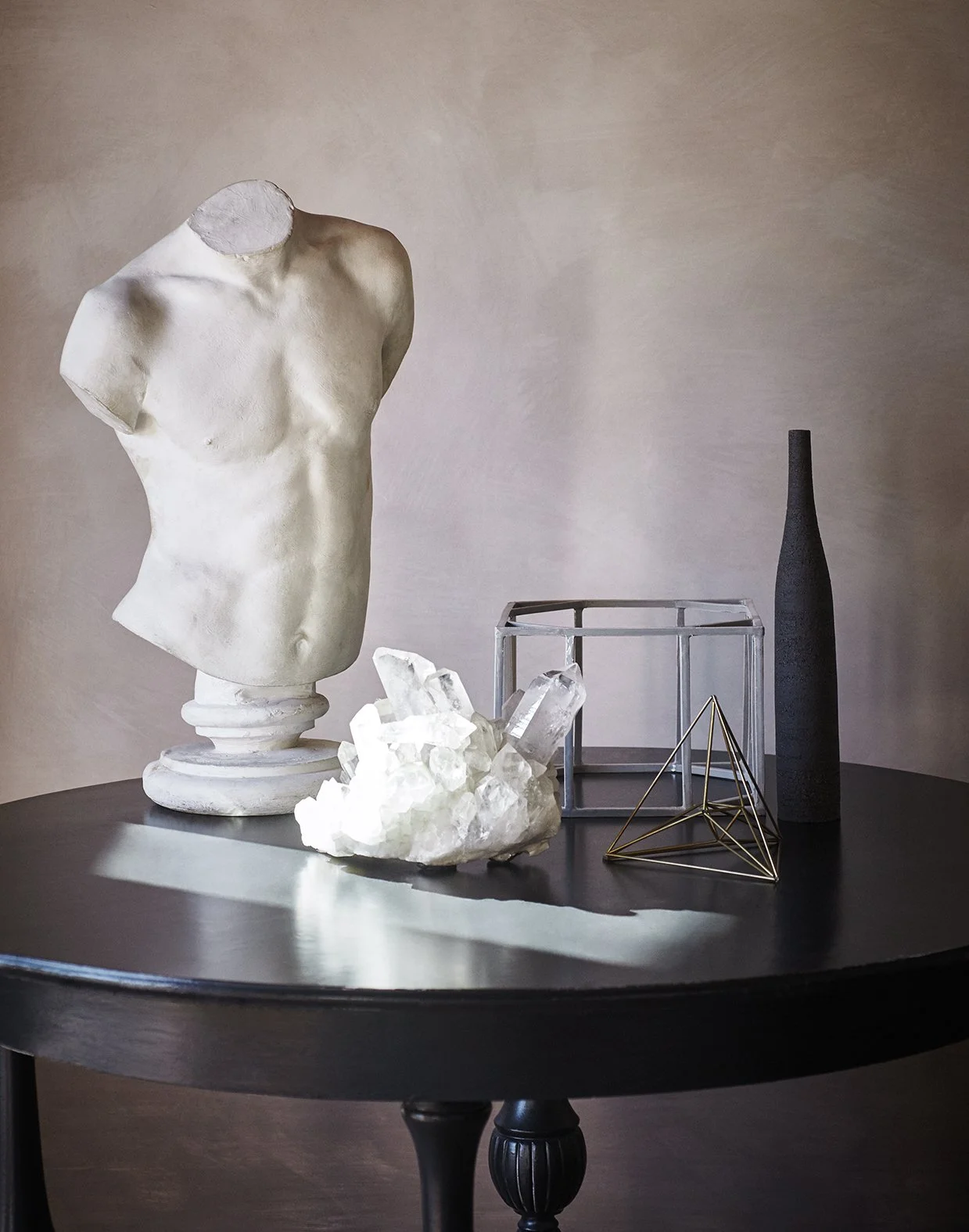 Eine schwarze Tisch mit mehreren Dekorationsgegenständen, darunter eine weiße antike Büste, ein Kristallcluster, eine offene Glasbox, eine schwarze Vase und eine goldene geometrische Skulptur, vor einer neutralen Wand.