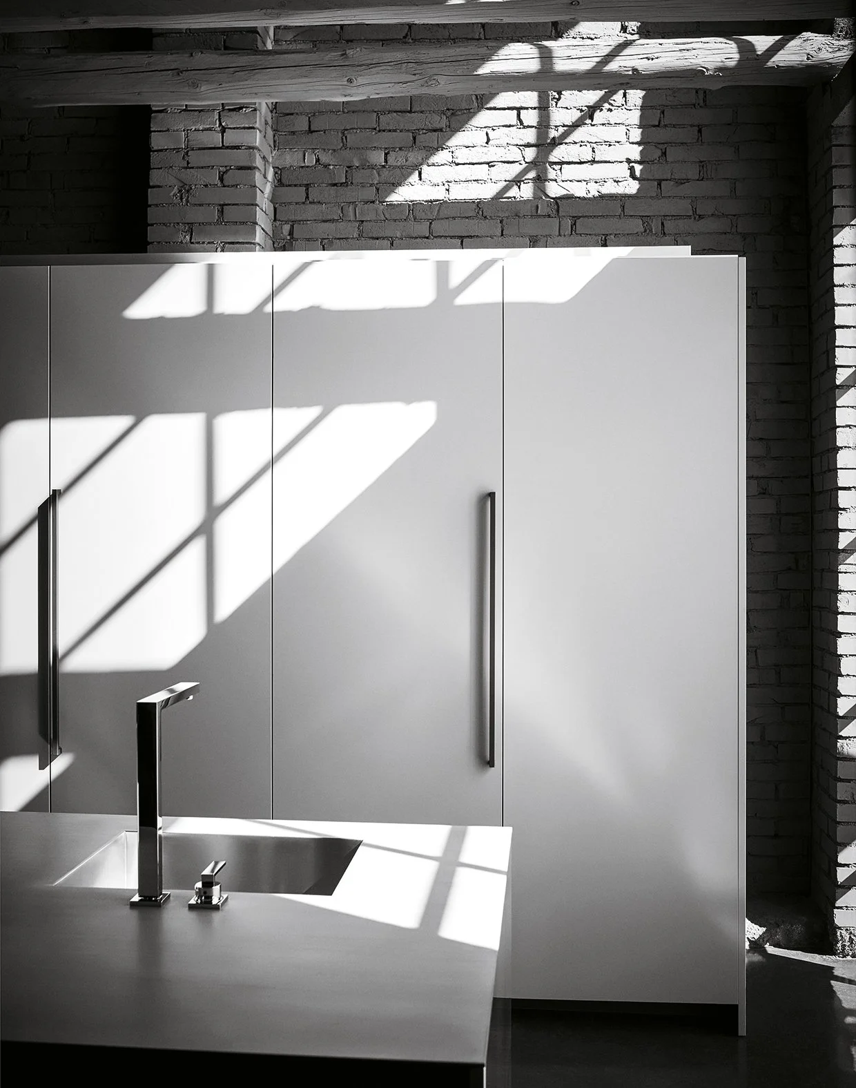 Moderne Küche mit weißen Schränken, einer Spüle, einem Wasserhahn und natürlichem Licht, das Schatten auf die Oberflächen wirft, vor einer Ziegelsteinwand.