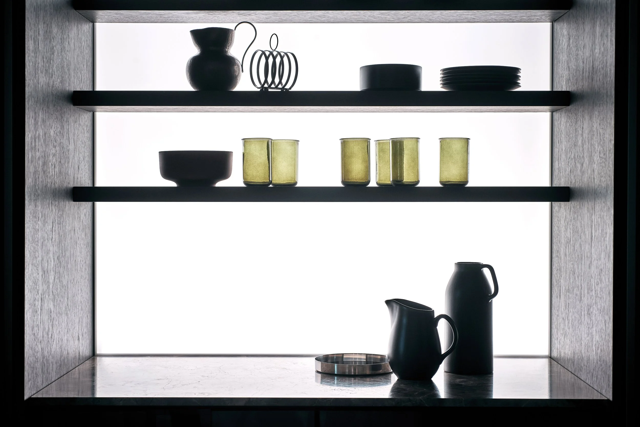Schwarze Schale, vier grüne Trinkgläser, schwarze Kaffeemaschine, Messbecher auf einer Küchenablage vor einem beleuchteten Hintergrund.