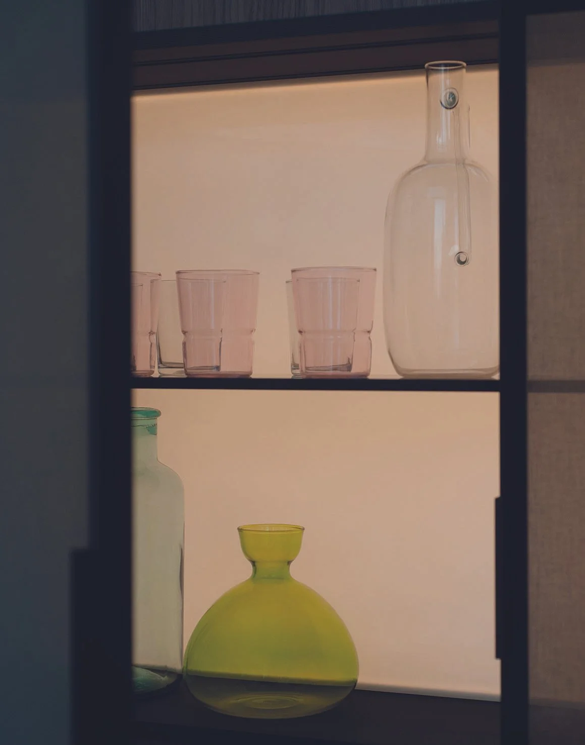 Blick in eine Vitrine mit vier pinkfarbenen Gläsern auf der oberen Ablage, einer weißen Flasche und einer gelben Vase auf der unteren Ablage.