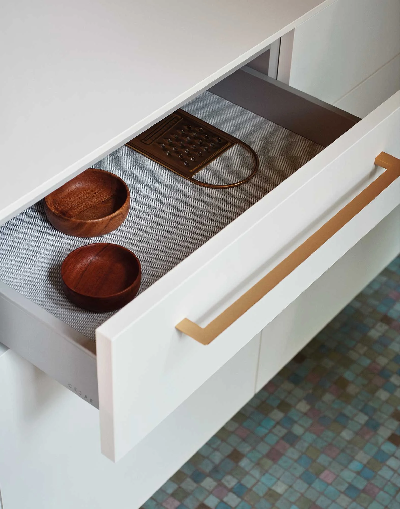 Ein weißer Schrank mit geöffneter Schublade, in der zwei kleine, runde Holzschalen und ein Holzbrett mit runden Metallknöpfen liegen. Der Boden ist mit bunten, kleinen Fliesen ausgelegt.