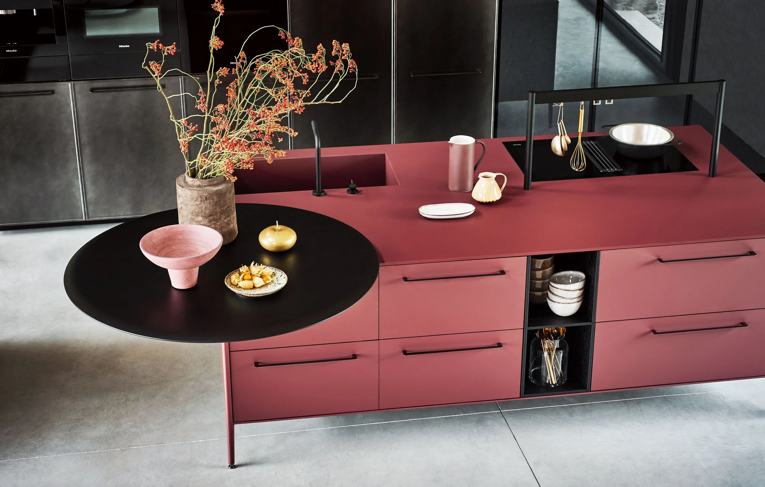 Moderne schwarze und rote Küchenschränke mit Küchenutensilien und Dekoration, darunter eine große Vase mit getrockneten roten Pflanzen, eine pinke Kaffeetasse, eine weiße Schüssel und andere Geschirrstücke.