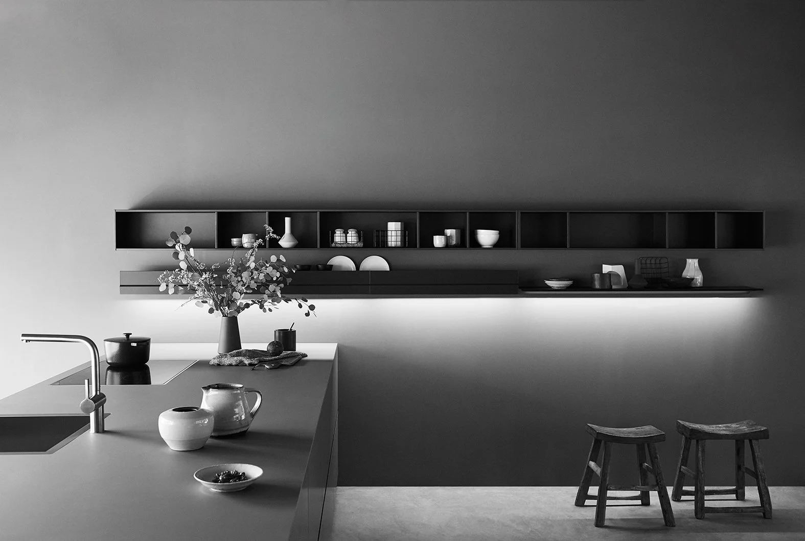 Moderne, minimalistische Küche mit dunklem Wandregal, weißen Geschirr und Dekoration, Holzbarhocker, Vasen, Küchenutensilien, und einem Küchenblock mit Wasserhahn, in Schwarz-Weiß-Darstellung.