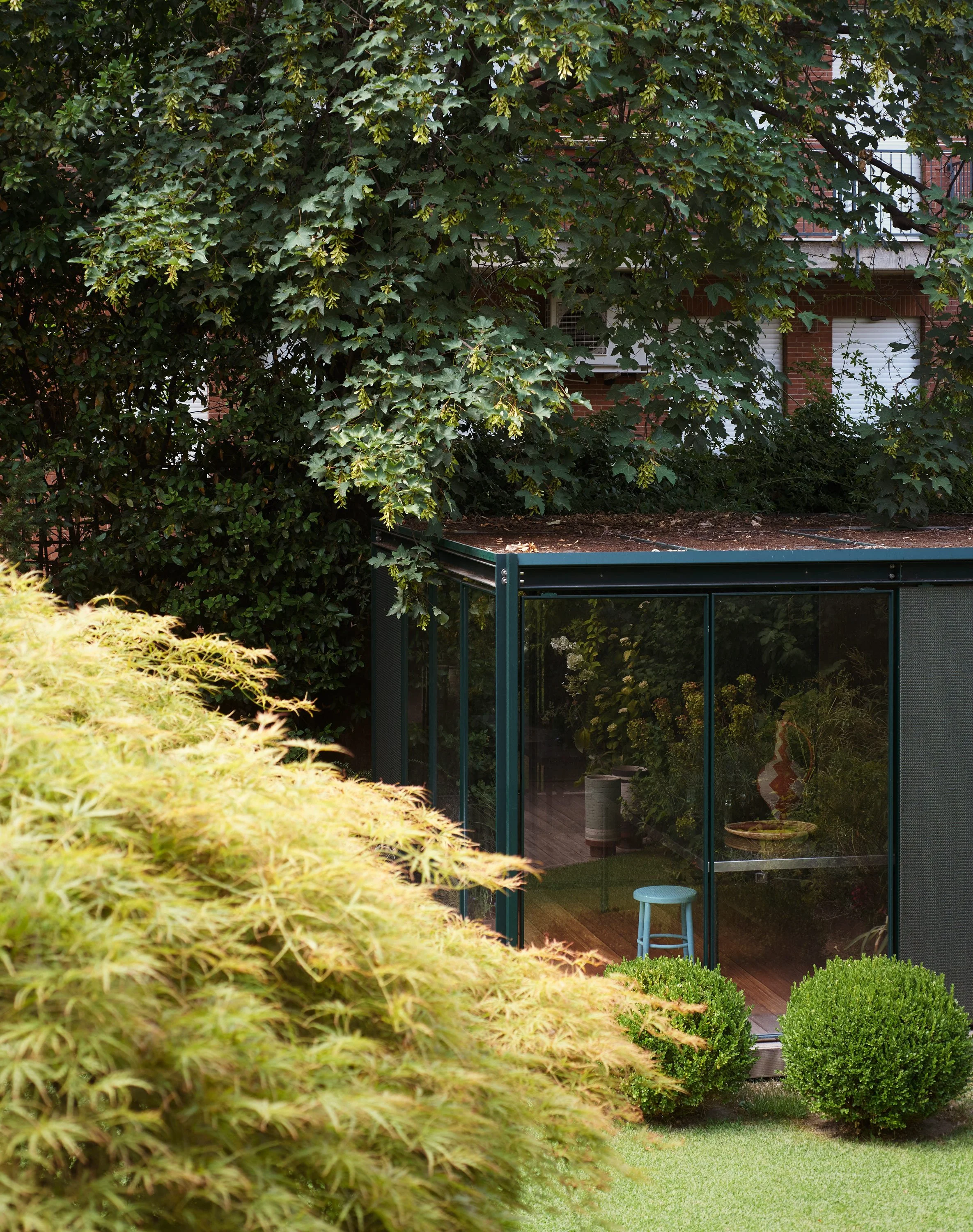 Garten mit grünem Rasen, Büschen und Bäumen sowie einem Wintergarten. Im Hintergrund ein Haus mit Balkon und Rollläden.
