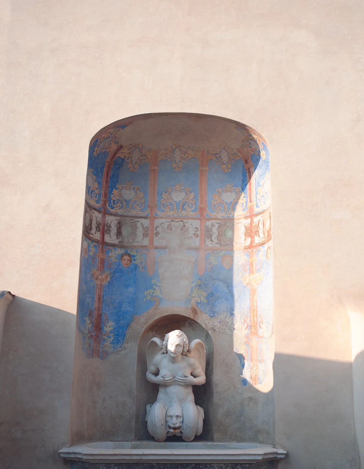 Eine Skulptur einer weinenden Figur, die von einer Löwenmähne und einem Kopf mit offenem Mund getragen wird, vor einer Wand mit einer farbenfrohen, dekorativen Malerei.