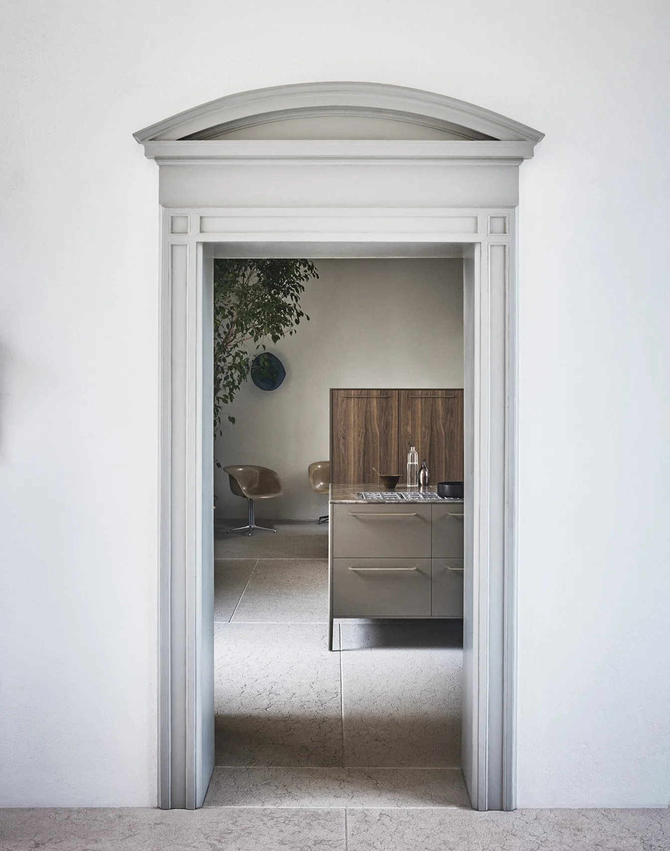 Blick durch eine Innentür in eine moderne Küche mit grauem Schrank, Holzabdeckung, Geschirr und Wasserkrügen, und hinter ihr ein Sitzbereich mit zwei Stühlen und Dekorationen wie einer Pflanze und Wandkunst.