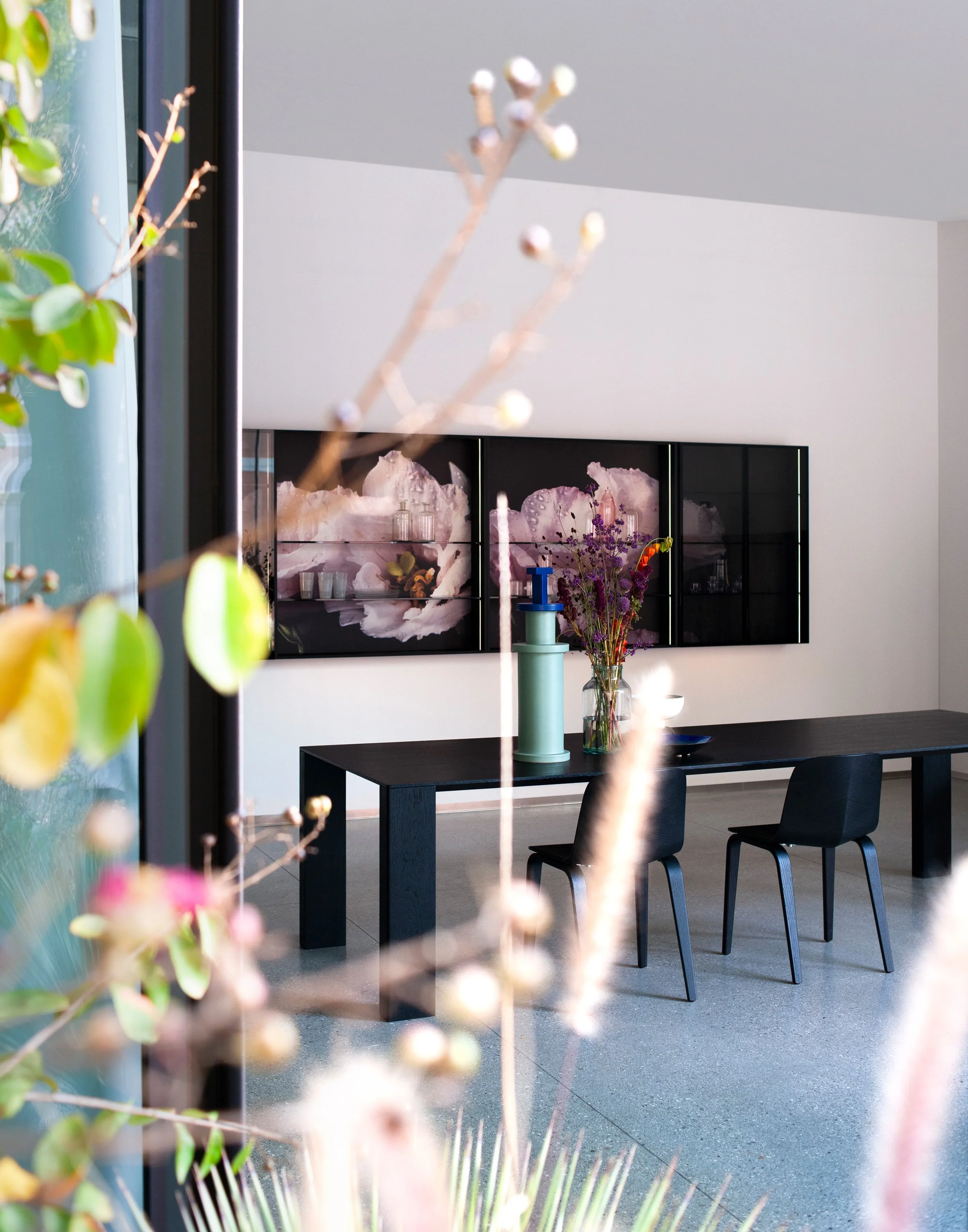 Innenaufnahme eines modernen Raums mit schwarzem Tisch, sechs schwarzen Stühlen, einer Wandmontage mit kunstvoll gestaltetem Fenster, einer Vase mit Blumen und einem schwarzen Sideboard mit Dekorationsartikeln.
