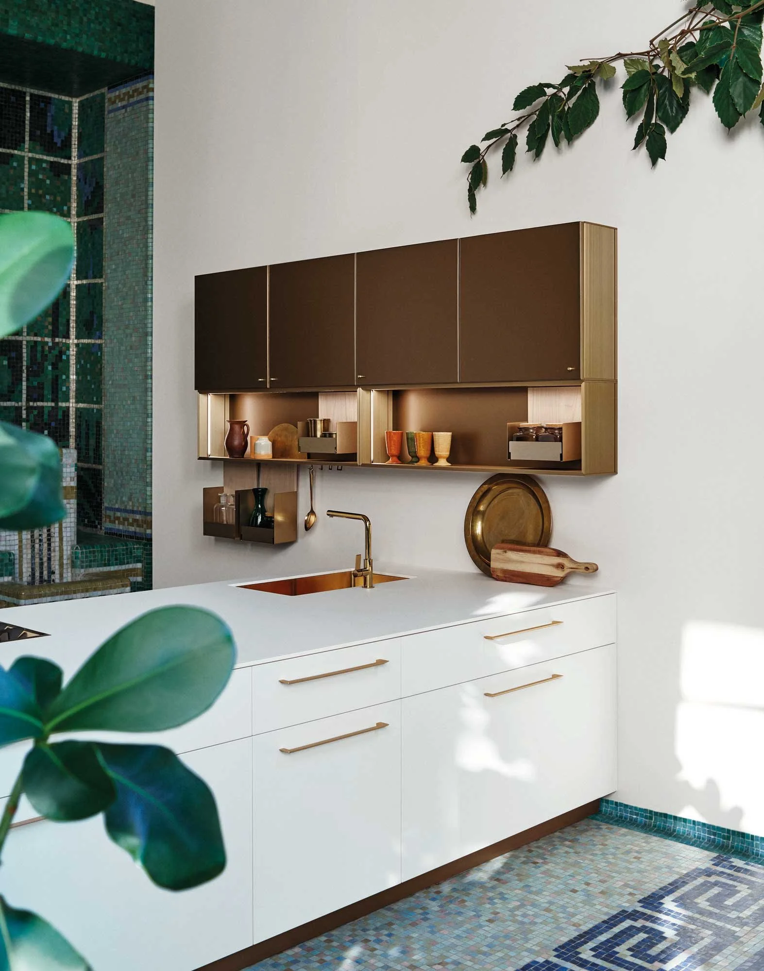 Moderne Küchenschrank mit weißen Türen und goldenen Griffen, offene Regale mit Geschirr und Dekor, kupferner Wasserhahn, Mosaikfliesenboden, grüne Zimmerpflanze im Vordergrund