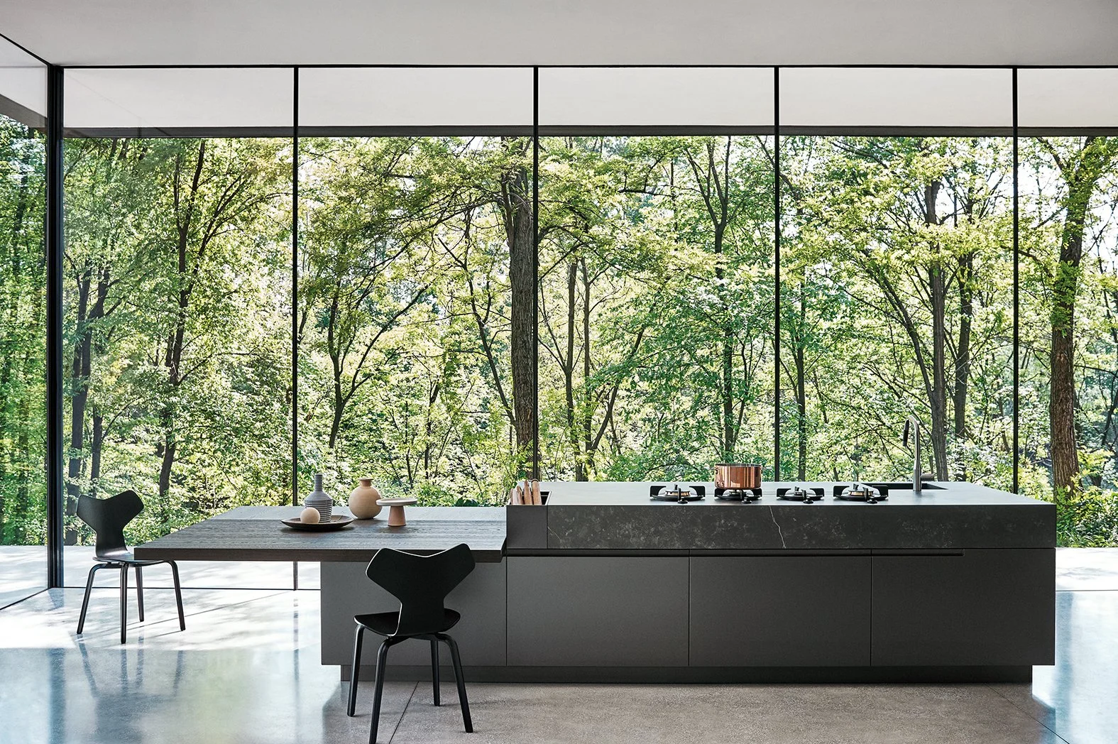 Moderne, minimalistische Küche mit großem Fenster, das eine grüne Waldlandschaft zeigt. Die Küche hat eine schwarze Insel mit Herd und Wasserhahn, eine separate Holzarbeitsplatte mit Dekorvasen und Tischstühlen.