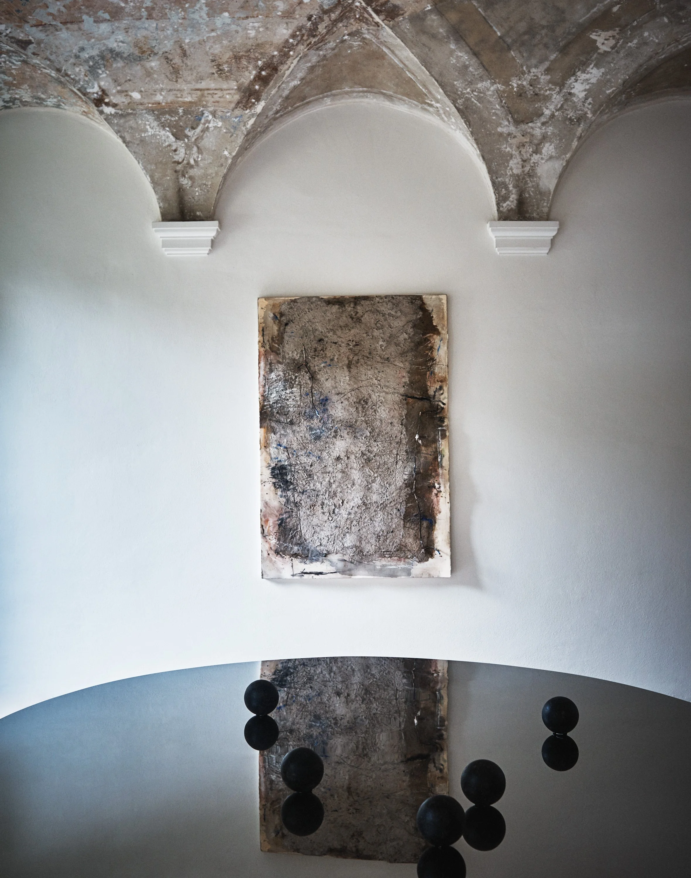 Eine Kunstinstallation in einem Raum mit weißer Wand und einem abgenutzten, bröckelnden Deckenhintergrund. Es gibt eine größere Leinwand oder Tafel mit abstrakter, erdiger und schwarzer Farbgebung, die an die Wand gelehnt ist. Im Vordergrund sind meh