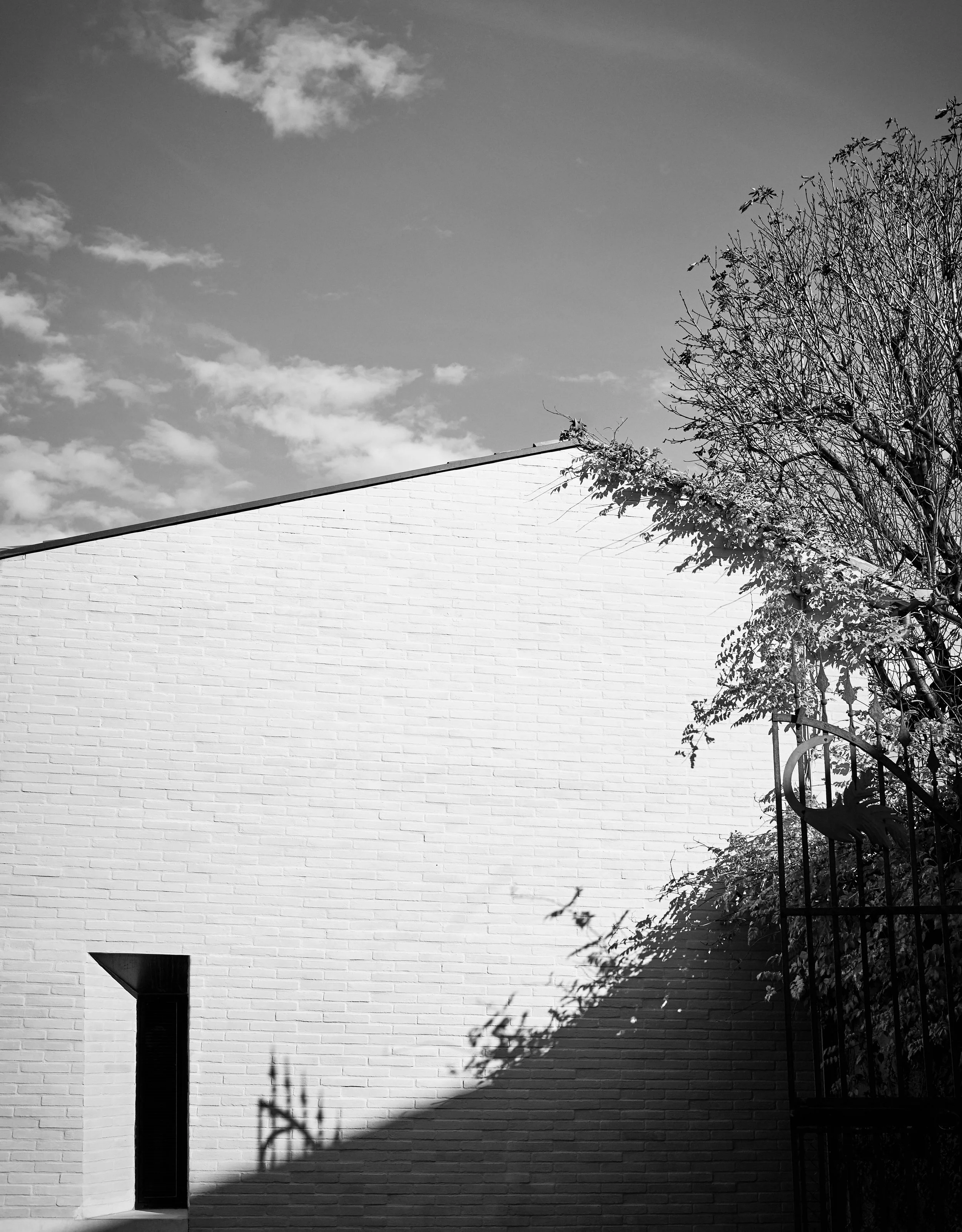 Schwarze und weiße Aufnahme eines Gebäudes mit einer weißen Backsteinmauer, einem kleinen oberirdischen Eingang, einem Fenster, einem Baum und einem Zaun, Schatten und Wolken am Himmel.