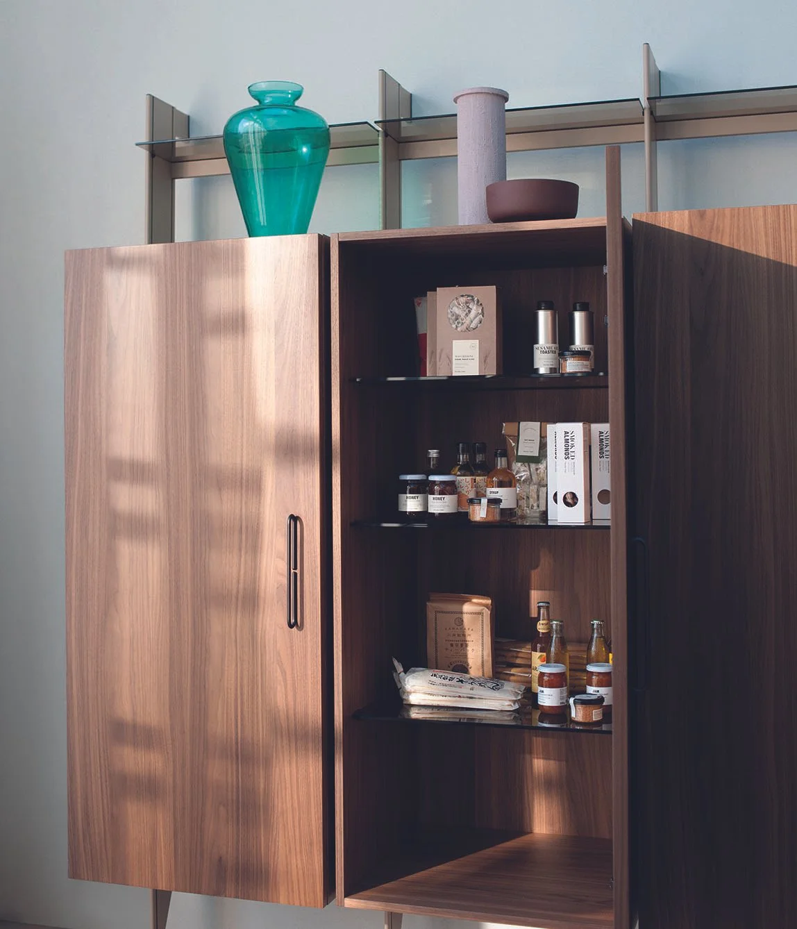 Offene Holzschrank mit Lebensmitteln und Küchenutensilien, dekorative Vase auf einem Regal oberhalb.