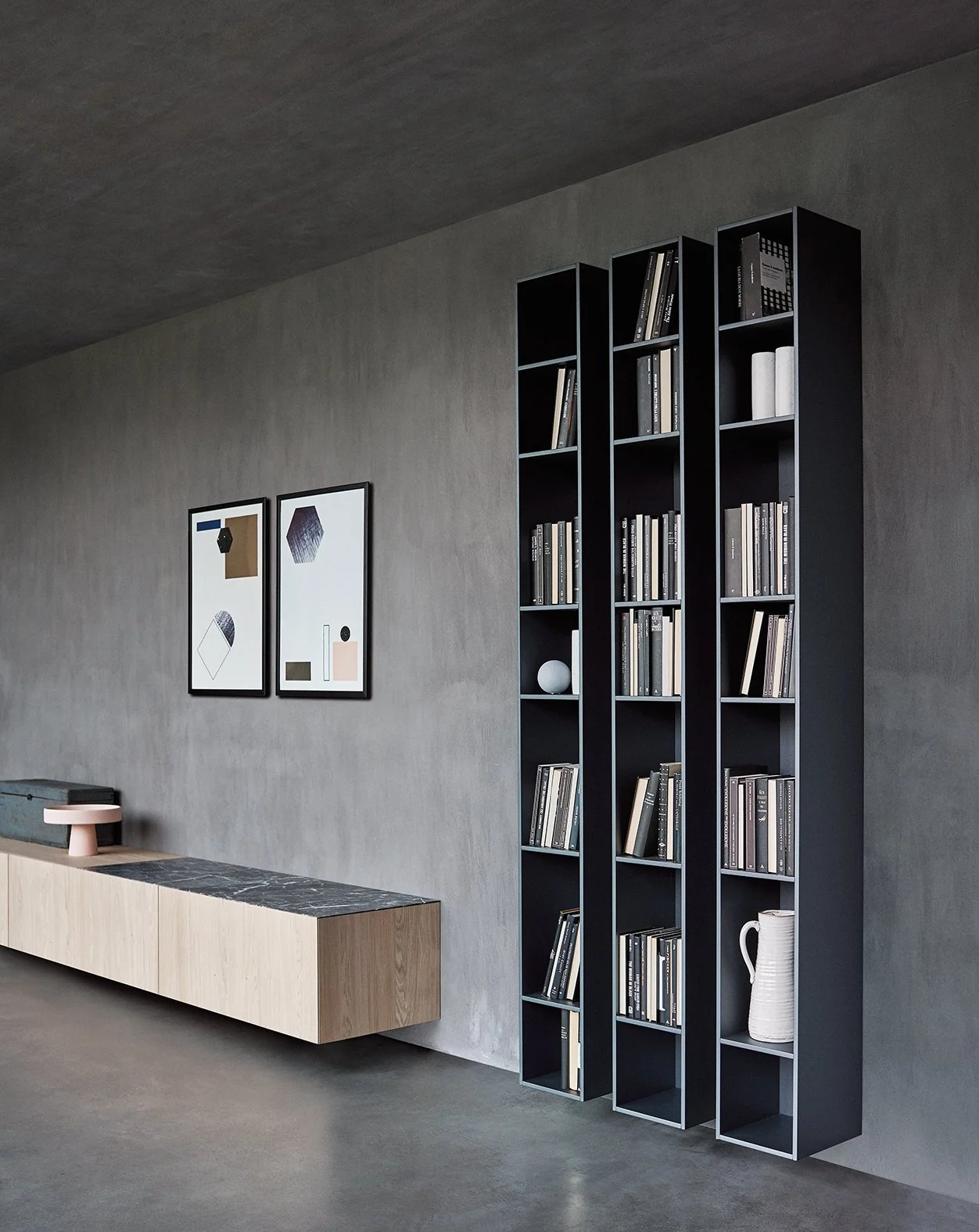 Moderner Raum mit grauer Wand und schwarzen Regalen, die Bücher und Dekorationsgegenstände enthalten. Zwei passende Bilder hängen an der Wand, und ein lowboard mit Dekorationsobjekten steht darunter.