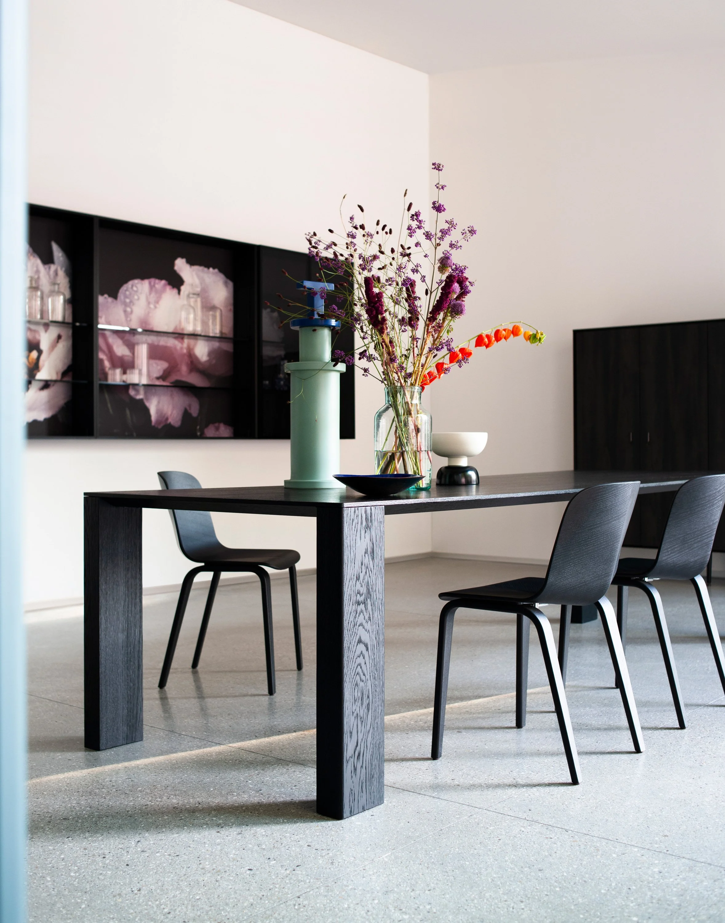 Moderner Esstisch mit schwarzem Holz mit Blumen in einer Glasvase, umgeben von schwarzen Stühlen in einem hellen, minimalistischen Raum.