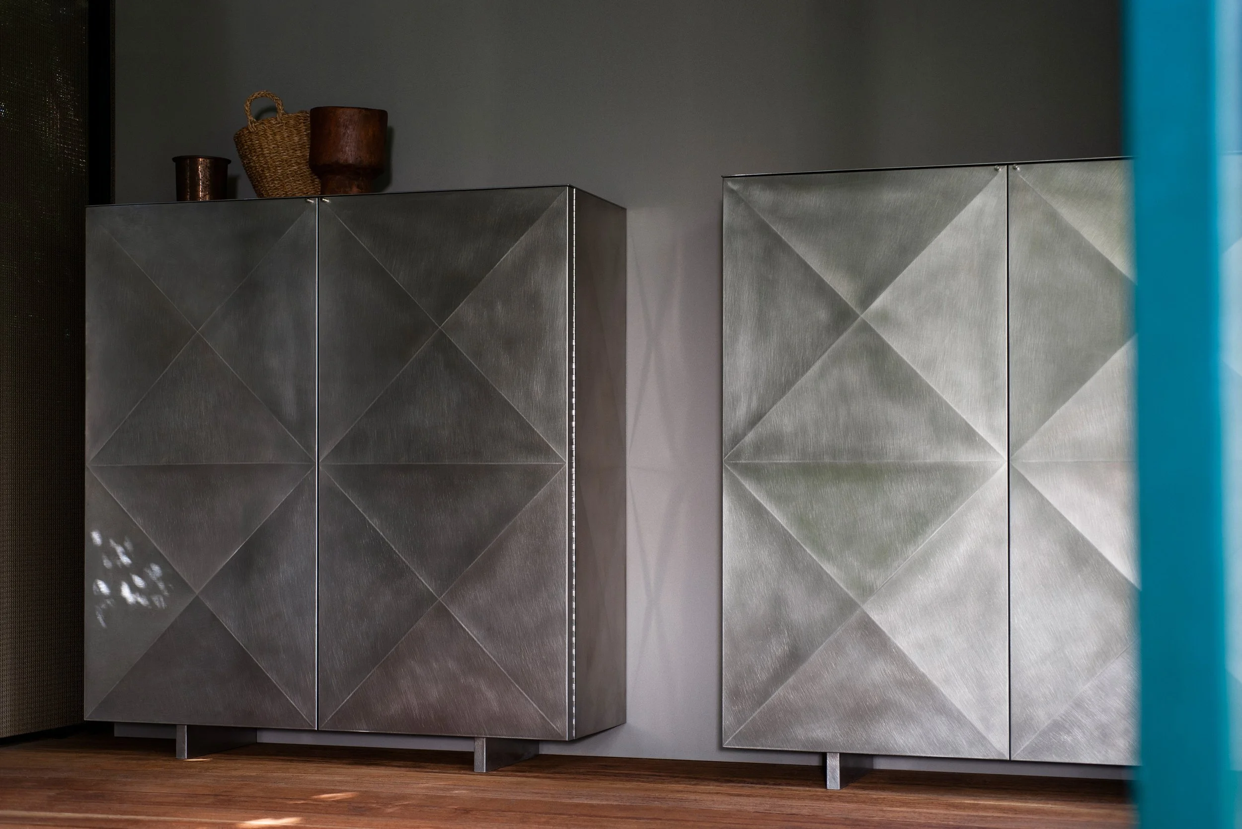 Moderner, metallicer Schrank in einem Raum mit grauer Wand und Holzboden, dekoriert mit drei Korb-ähnlichen Gegenständen auf dem Schrank.