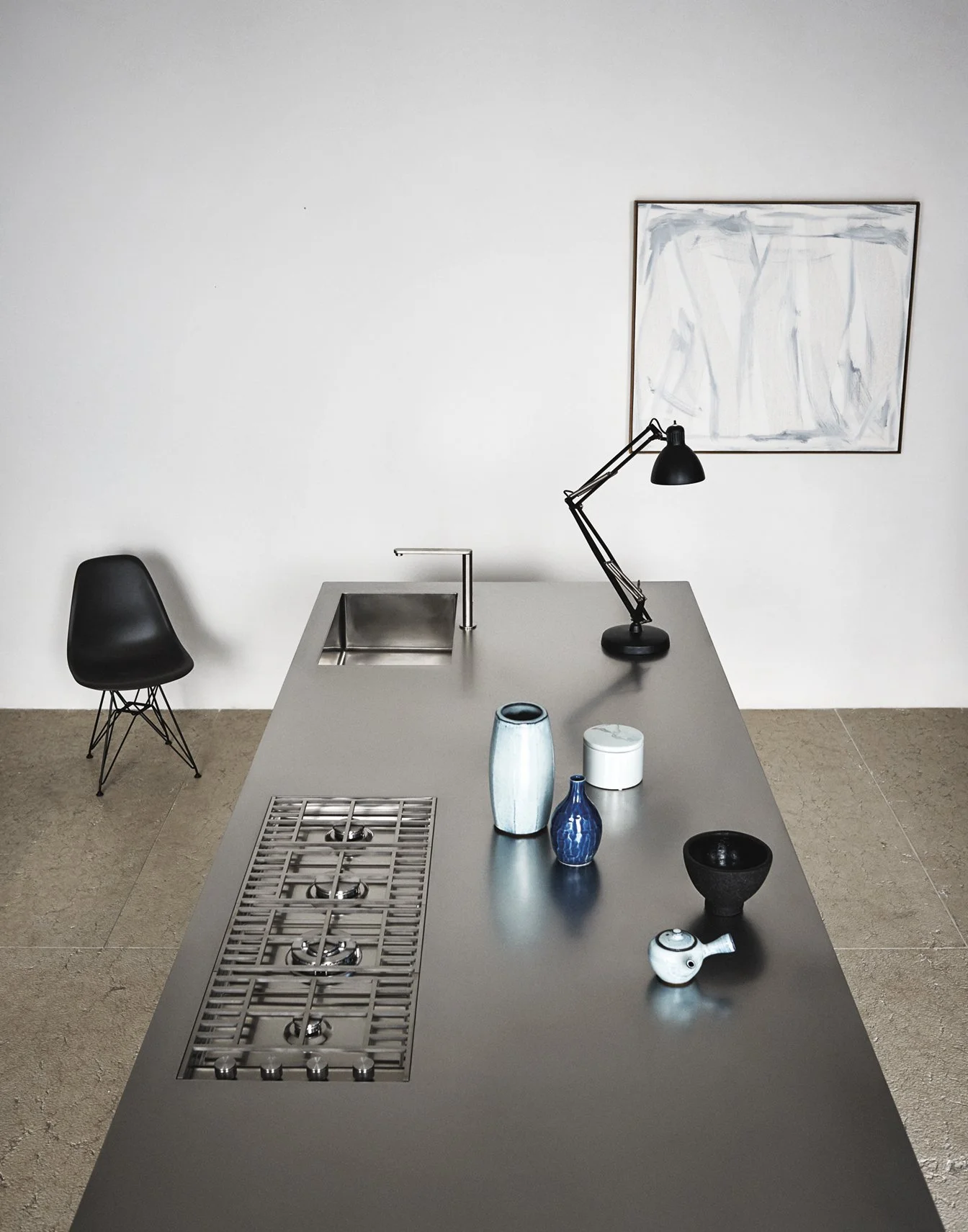 Moderne Küche mit grauem Küchenblock, schwarzen Stühlen, dekorativen Vasen und einer Tischlampe, im Hintergrund eine weiße Wand mit abstrakter Kunst.