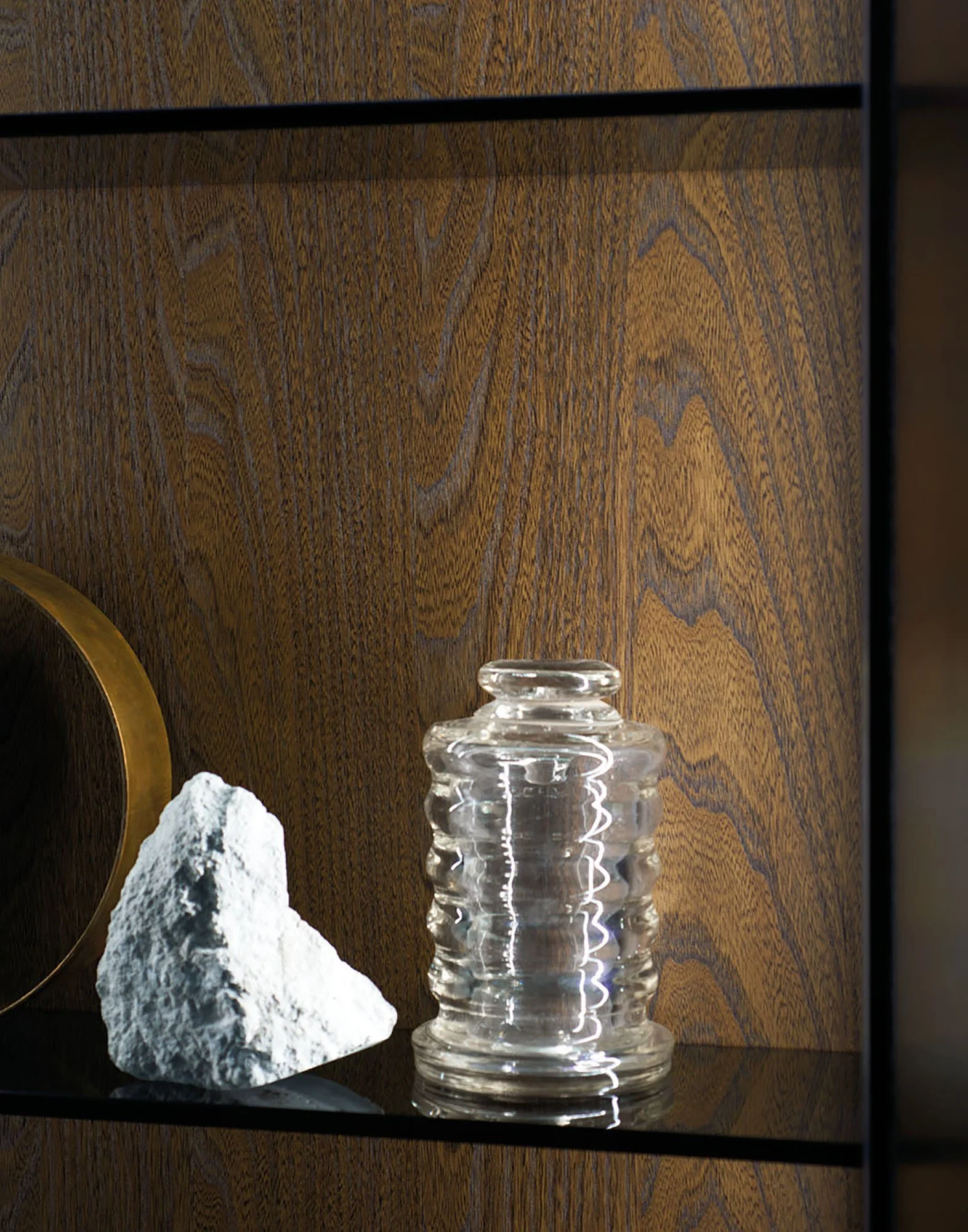 Ein Holzregal mit einer Glaskaraffe, einem weißen Stein und einem schwarzen Gegenstand im Hintergrund.