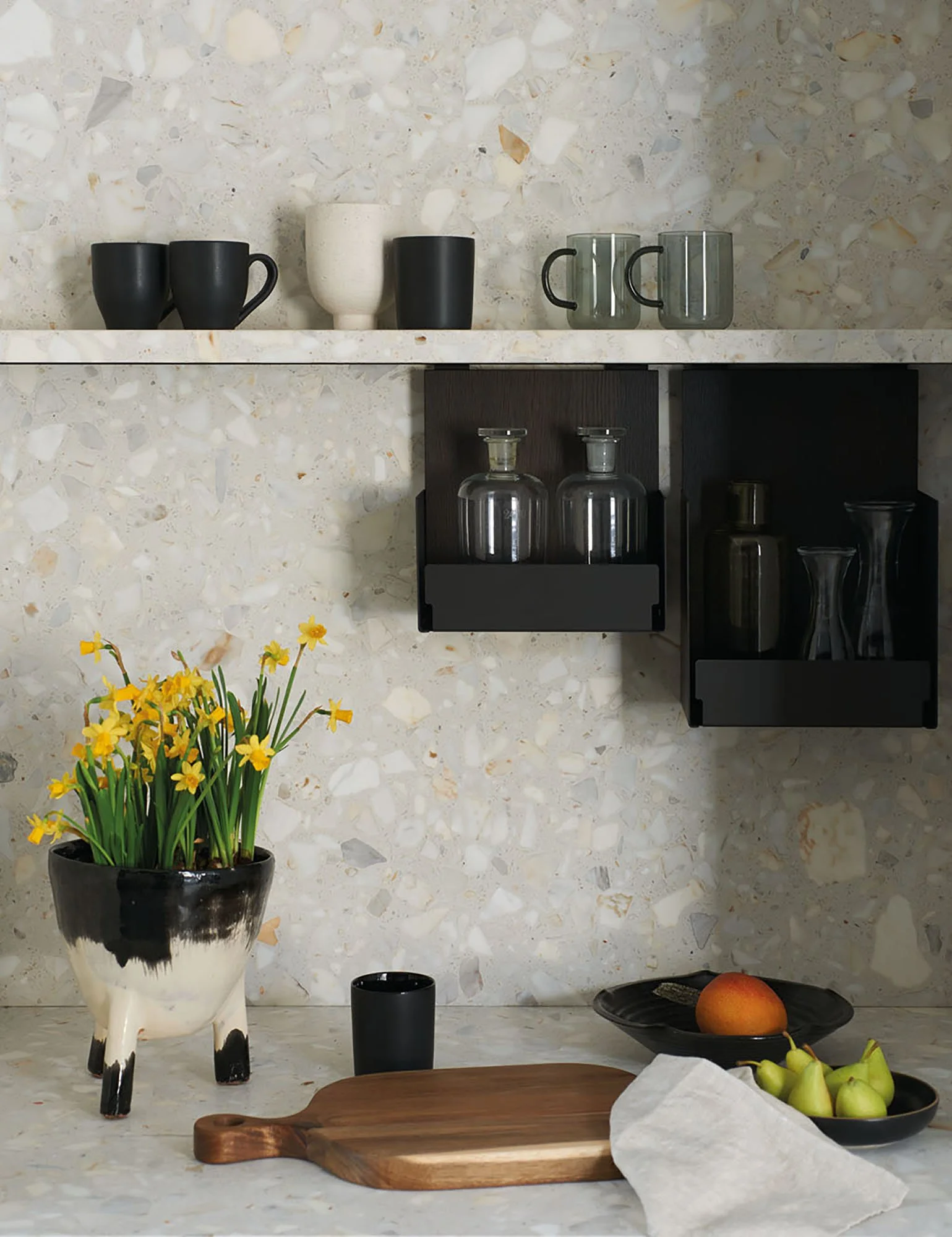 Ein Küchentresen mit schwarzen, weißen und grauen Tassen, Gläsern, Kannen und einer schwarzen Vase mit gelben Blumen, sowie einer Holzplatte mit Äpfeln und Birnen, und einer schwarzen Schale. Der Hintergrund besteht aus einer Marmorwand.