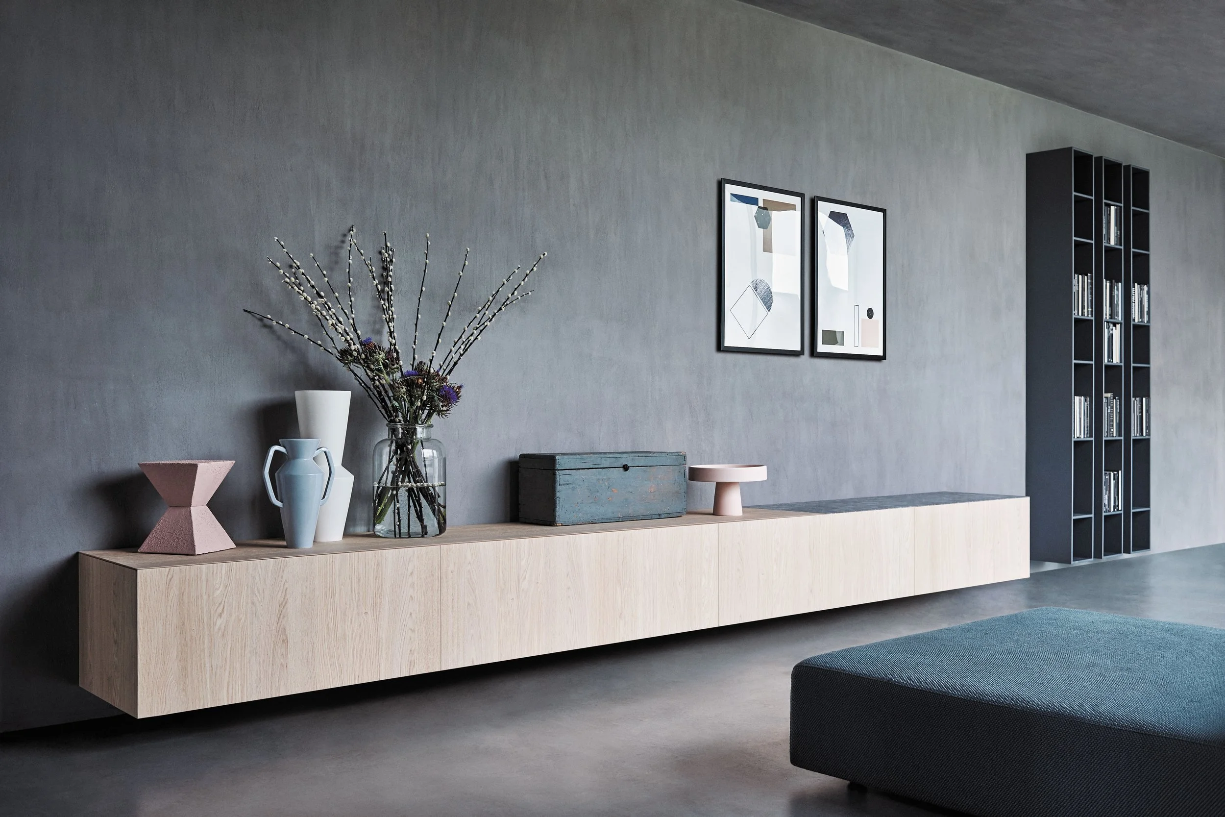 Ein moderner Wohnzimmerbereich mit einer langen hölzernen Sideboard, darauf Blumen in einer Glasvase, geometrische Dekorationen und zwei Bilder an der Wand, ein schwarzes Bücherregal an der rechten Seite.