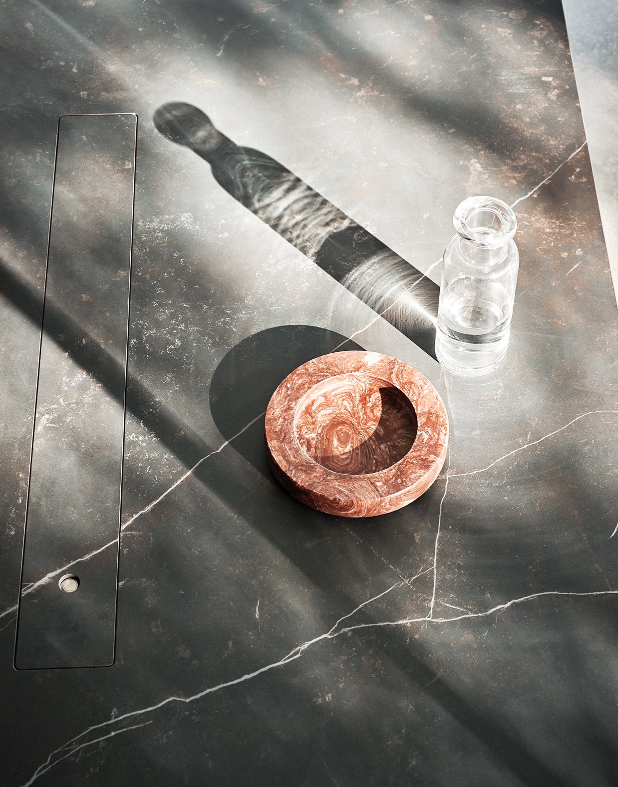 Auf einem dunklen Marmor-Tisch steht eine kleine Glasflasche mit Wasser, eine runde, rosafarben marmorierte Schale, die im Schatten liegt, und eine diagonale Schattenlinie, die durch die Szene läuft.
